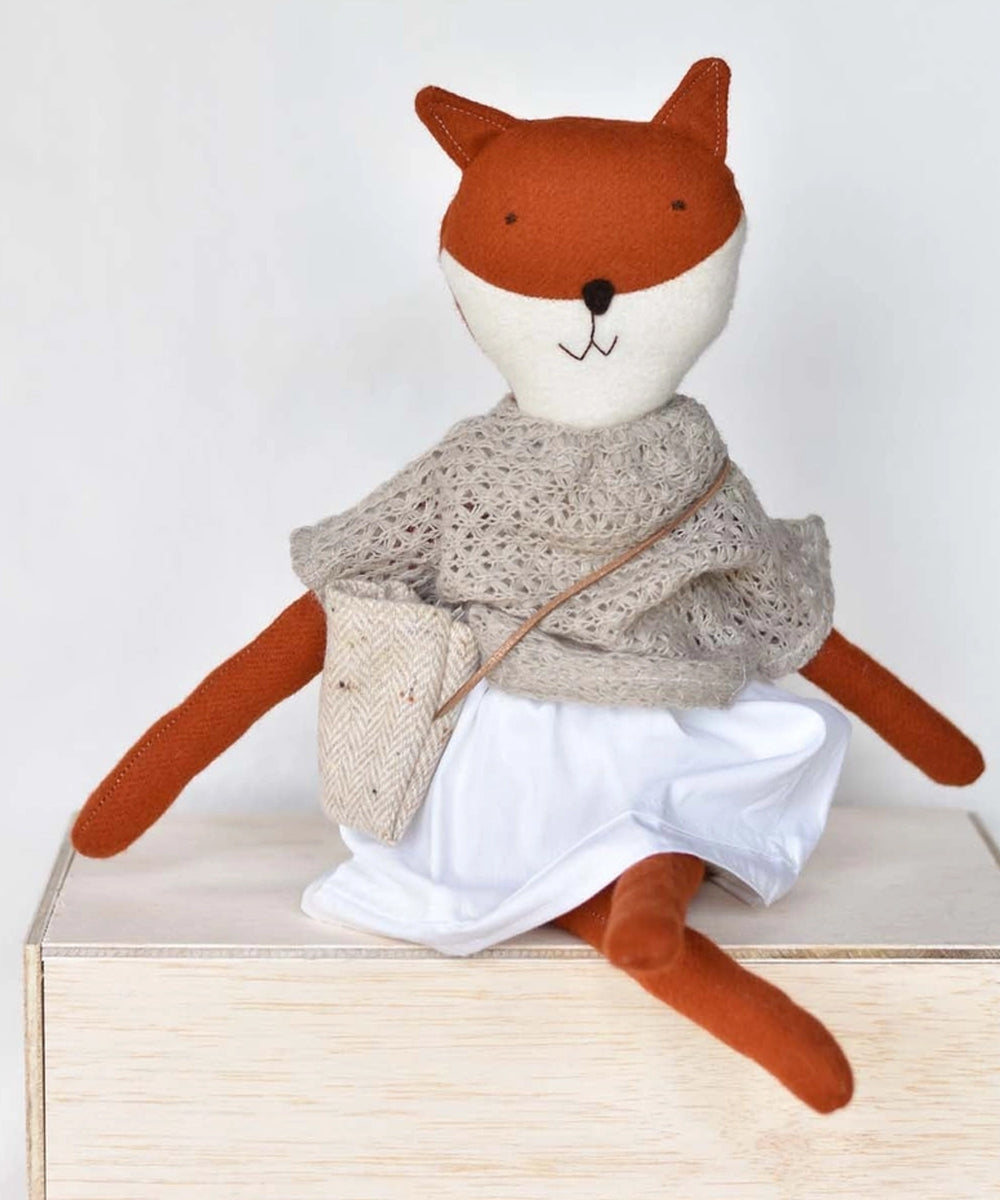 Florette le Fox doll in a white skirt, knitted shawl, and crossbody bag sits on a wooden surface.