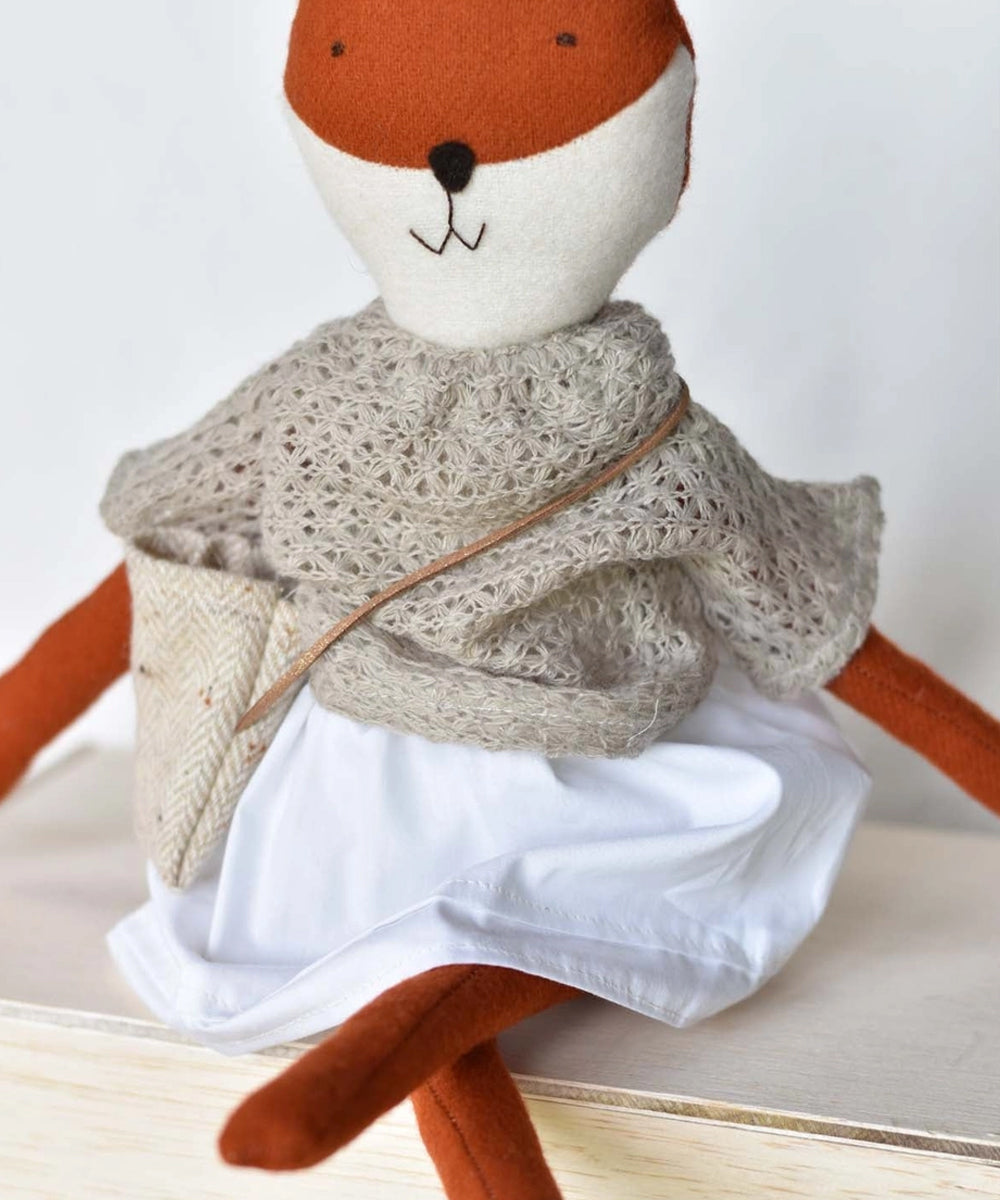 Florette le Fox, a stuffed fox doll in a knitted shawl, white skirt, and crossbody bag, sits on a wooden surface.