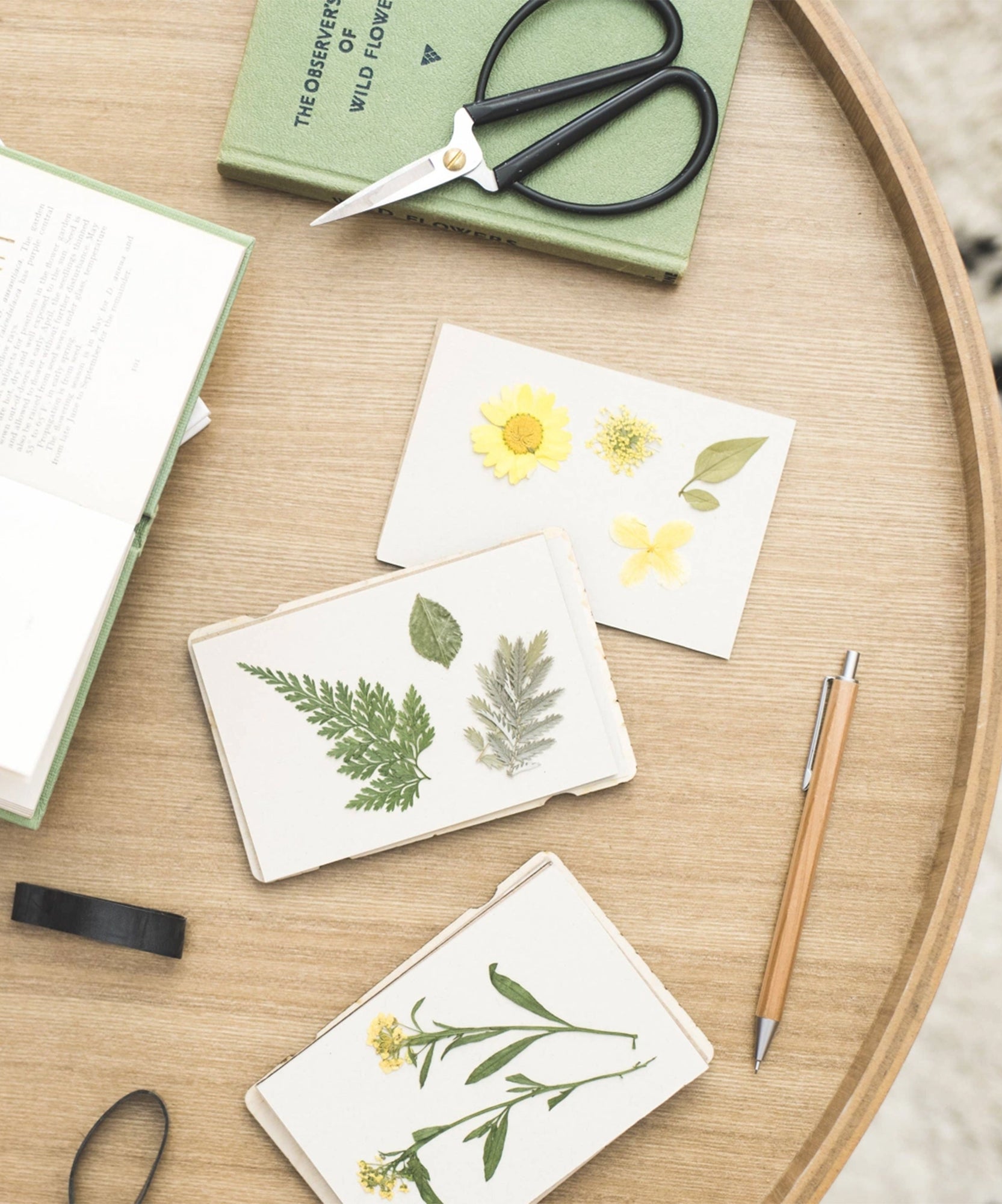 A round wooden table featuring a Flower Press, botanical cards, scissors, a book, a pen, and black tape.