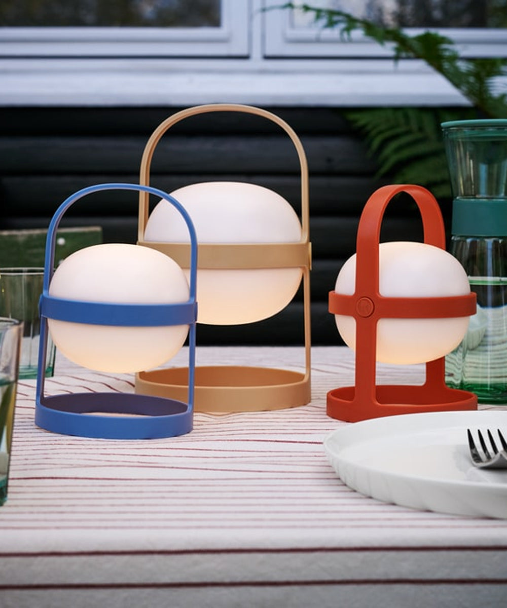 A Rosendahl Soft Spot Solar Circular, Sky Blue lantern sits outdoors on a striped tablecloth beside glassware and plates.
