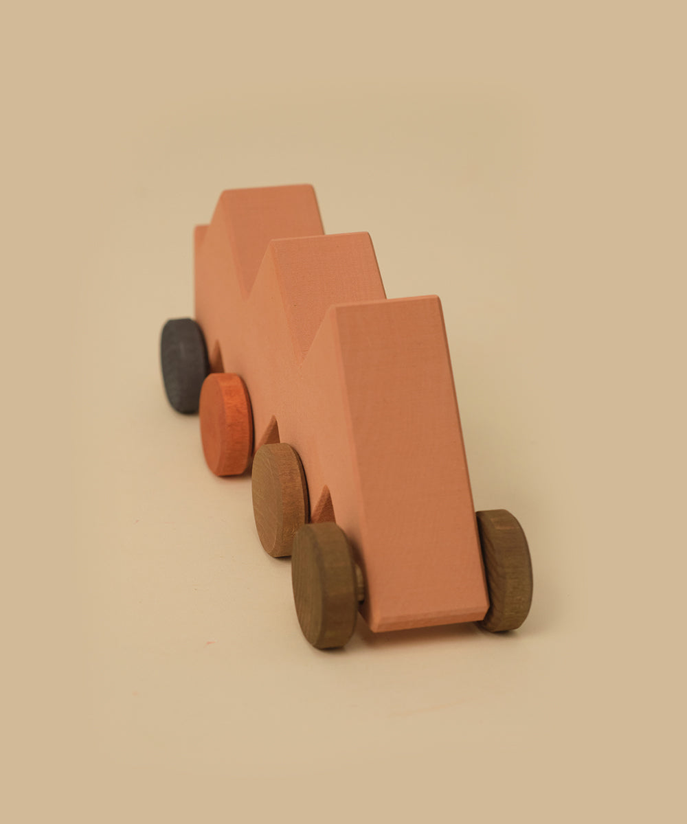 Le Zigzag Vehicle is a wooden toy car with six wheels, a zigzag-shaped top, and earth-tone colors on a beige background.