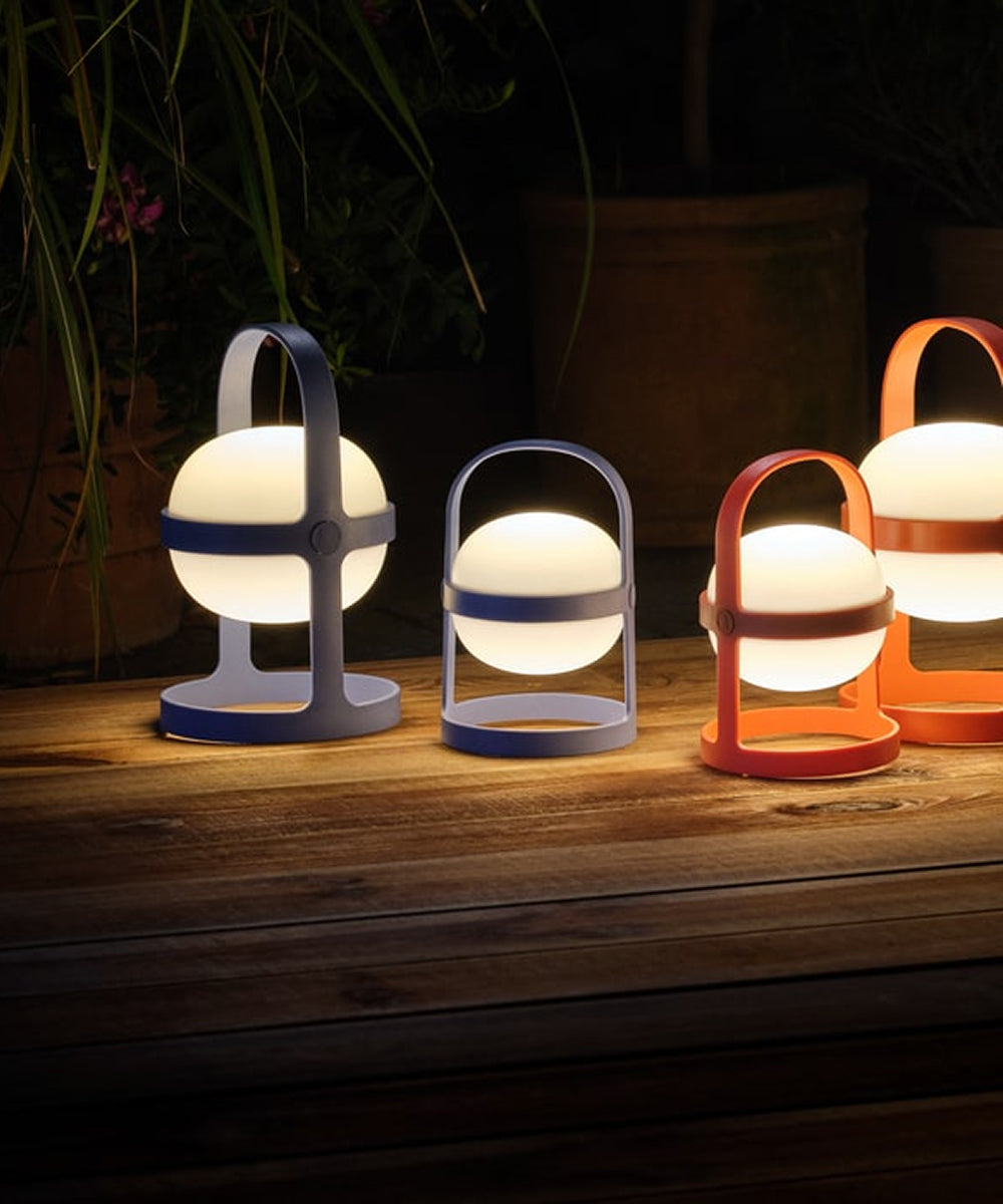 Four Rosendahl Soft Spot Solar Circular lanterns, two in sky blue, glow warmly with handles on a wooden surface outside at night.