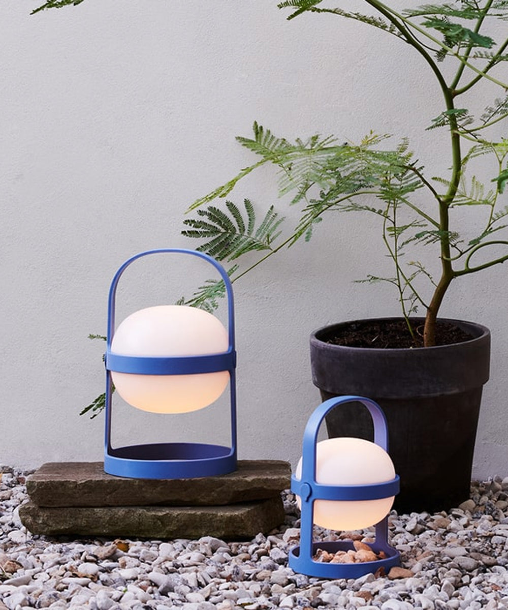 Two Rosendahl Soft Spot Solar Circular lamps in Sky Blue with round white lights sit on stones beside a green potted plant, against a gray wall.