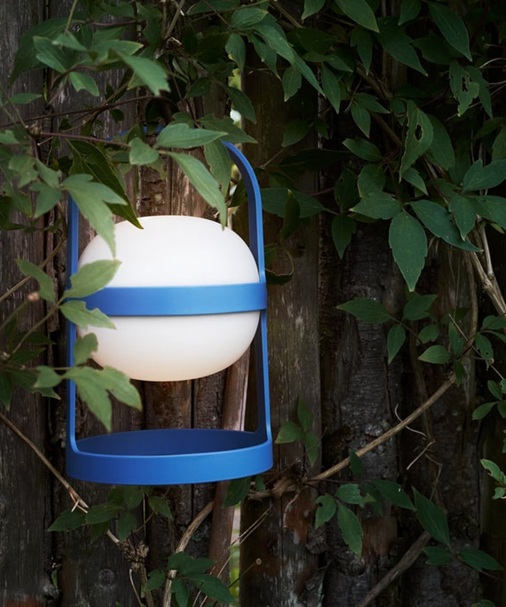 A Rosendahl Soft Spot Solar Circular in Sky Blue hangs on a wooden fence, framed by green leafy branches.
