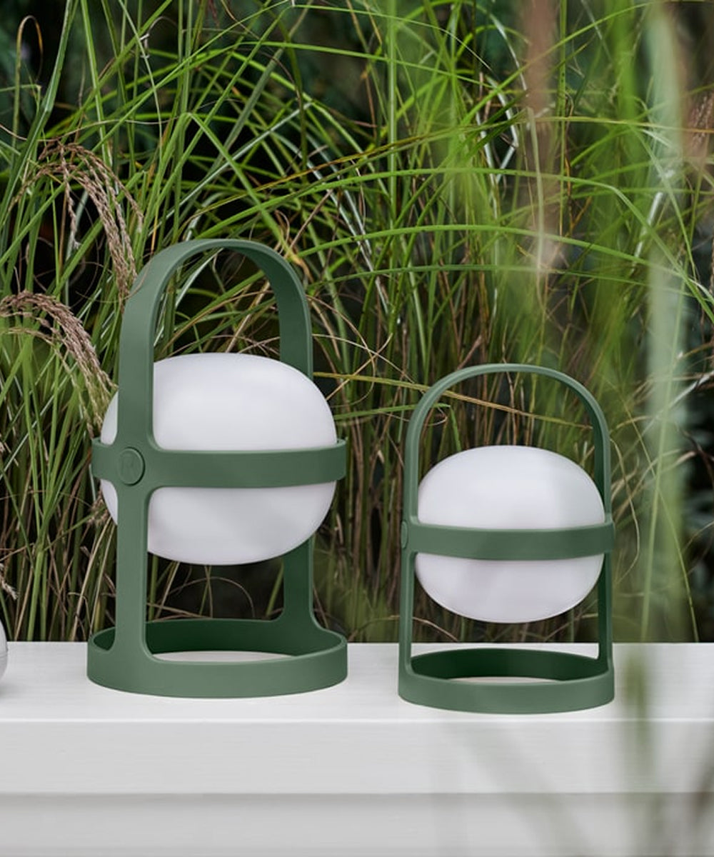 Two Rosendahl Soft Spot Solar Circular lanterns in pine green with white spheres sit on a white surface outdoors amid tall grass and plants.
