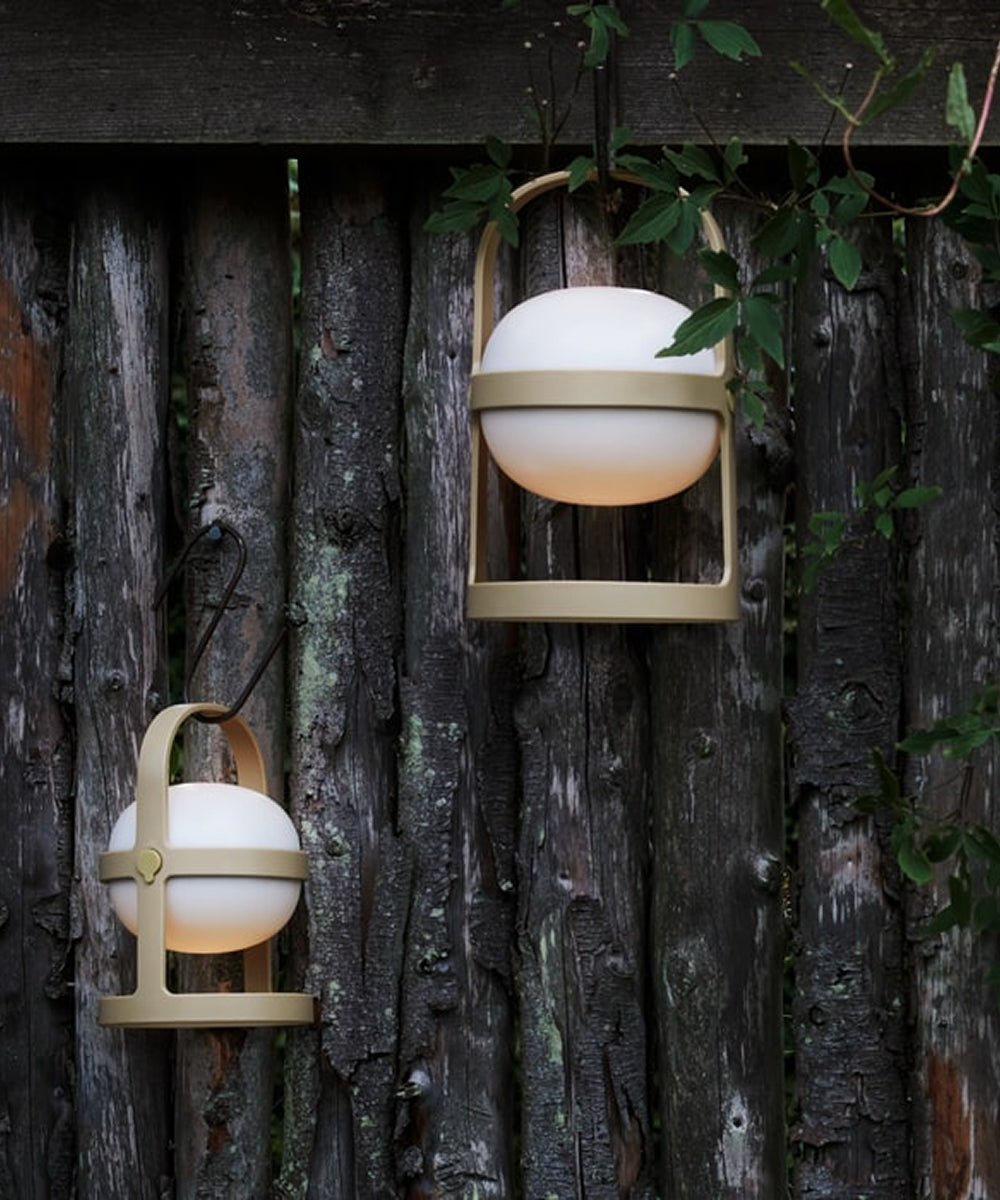 Two Rosendahl Soft Spot Solar Circular lanterns in wheat hang on weathered wood, round white lights glowing amid green leaves.