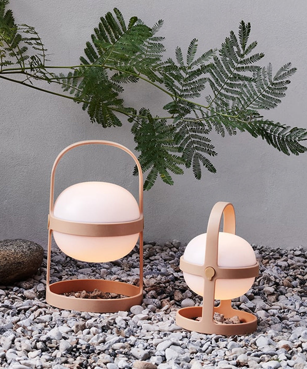 Two Rosendahl Soft Spot Solar Circular lanterns in Wheat with tan handles rest on pebbles, set against a wall and fern-like greenery.