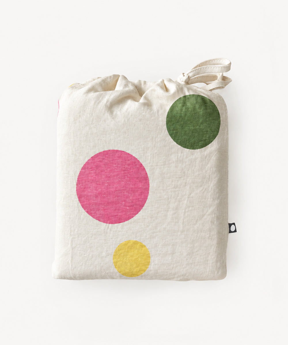 Linen Crib Sheet in folded cream fabric with a drawstring top and large pink, green, and yellow polka dots.