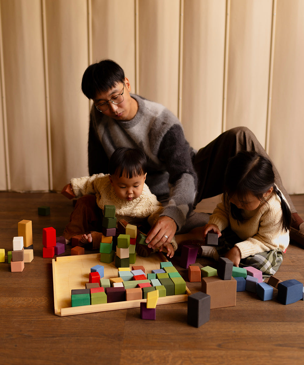 An adult and two children play indoors with Le Oddy Cubes, colorful wooden blocks, on a wooden floor.
