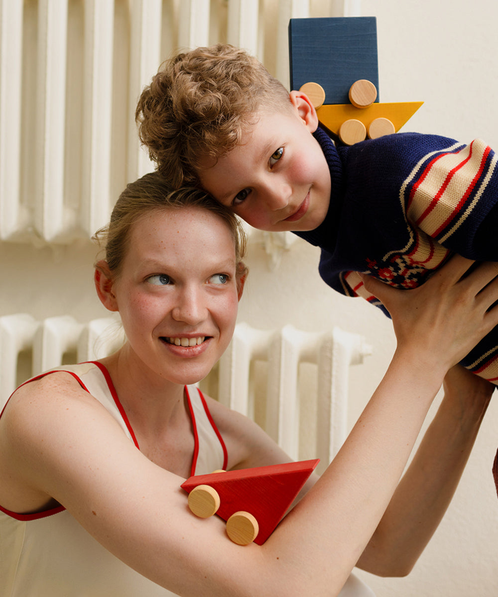 A woman lifts a smiling child holding Le Vehicle Trio wooden blocks, both looking happy and playful indoors.