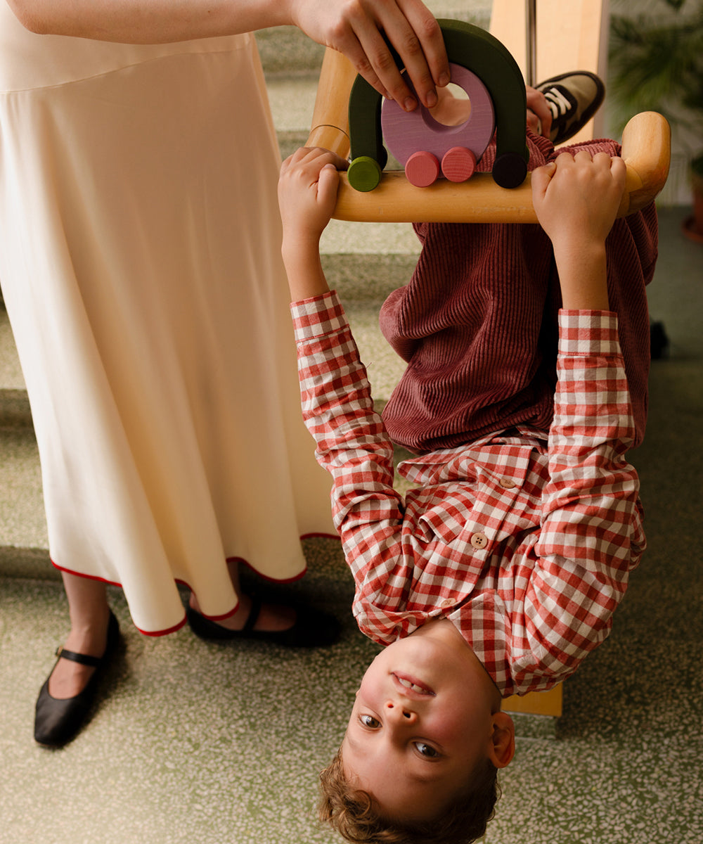 A smiling child hangs upside down from a chair, holding the Le Vehicle Duet toy, with an adult standing nearby.