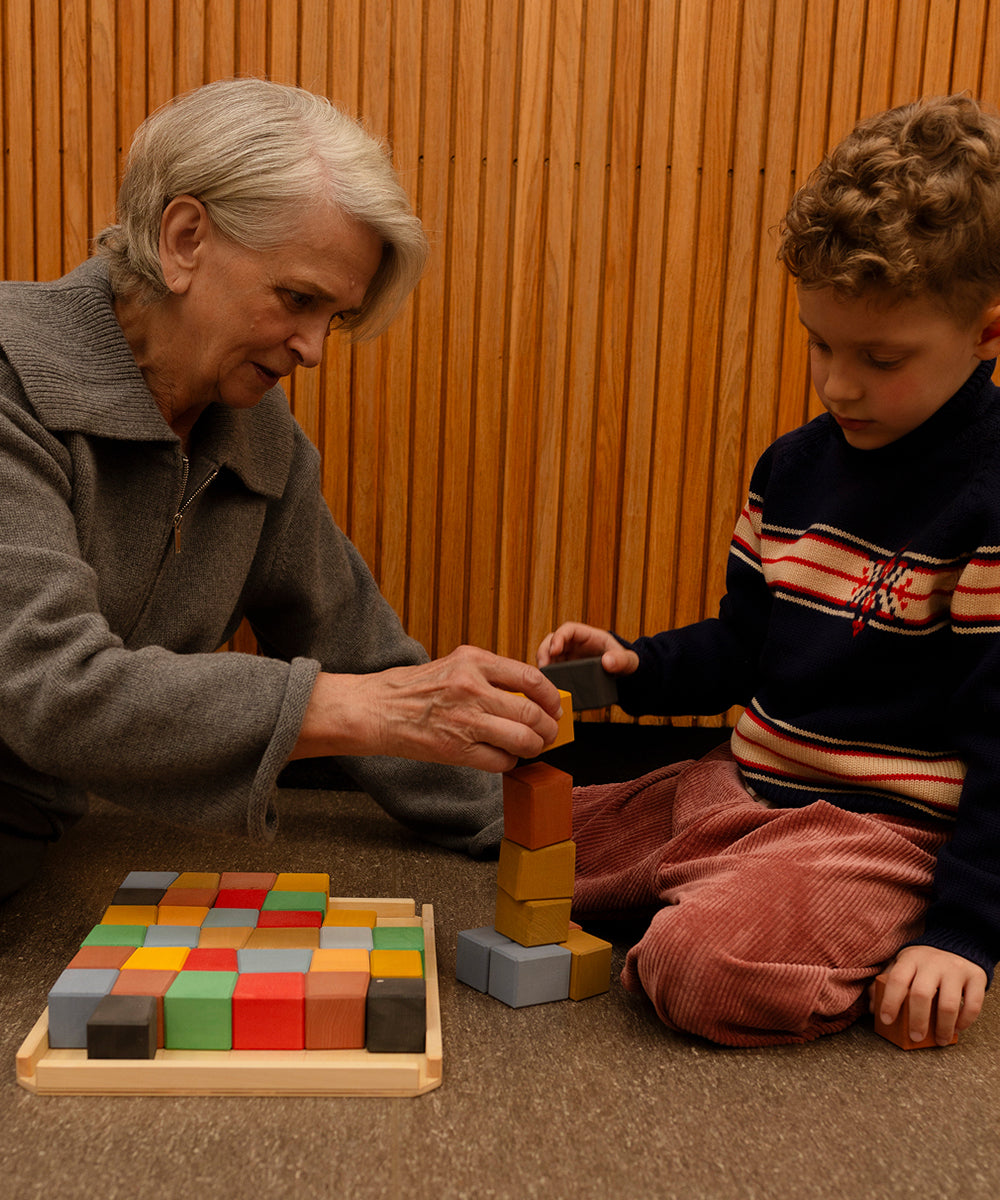 An elderly woman and a young boy stack Le Oddy Cubes - Mini together on the floor beside a wooden wall.