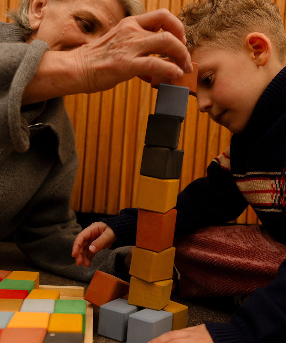 An older woman and a young boy stack Le Oddy Cubes - Mini, colorful wooden blocks, together on the floor.