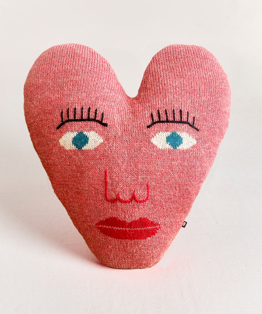 The Heart Pillow is heart-shaped with a knitted face, blue eyes, lashes, nose, and red lips on a pink background.