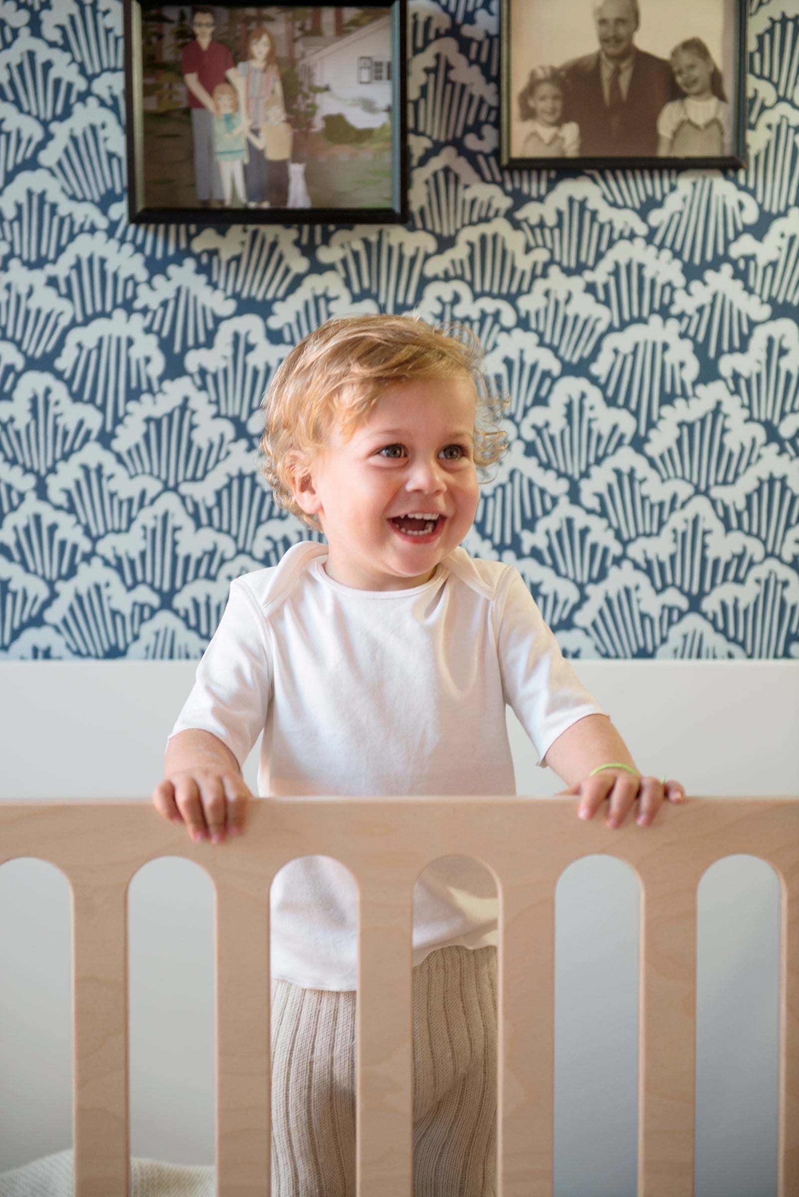 A toddler with curly hair stands smiling in a wooden crib, set against a blue patterned wall with framed photos above.