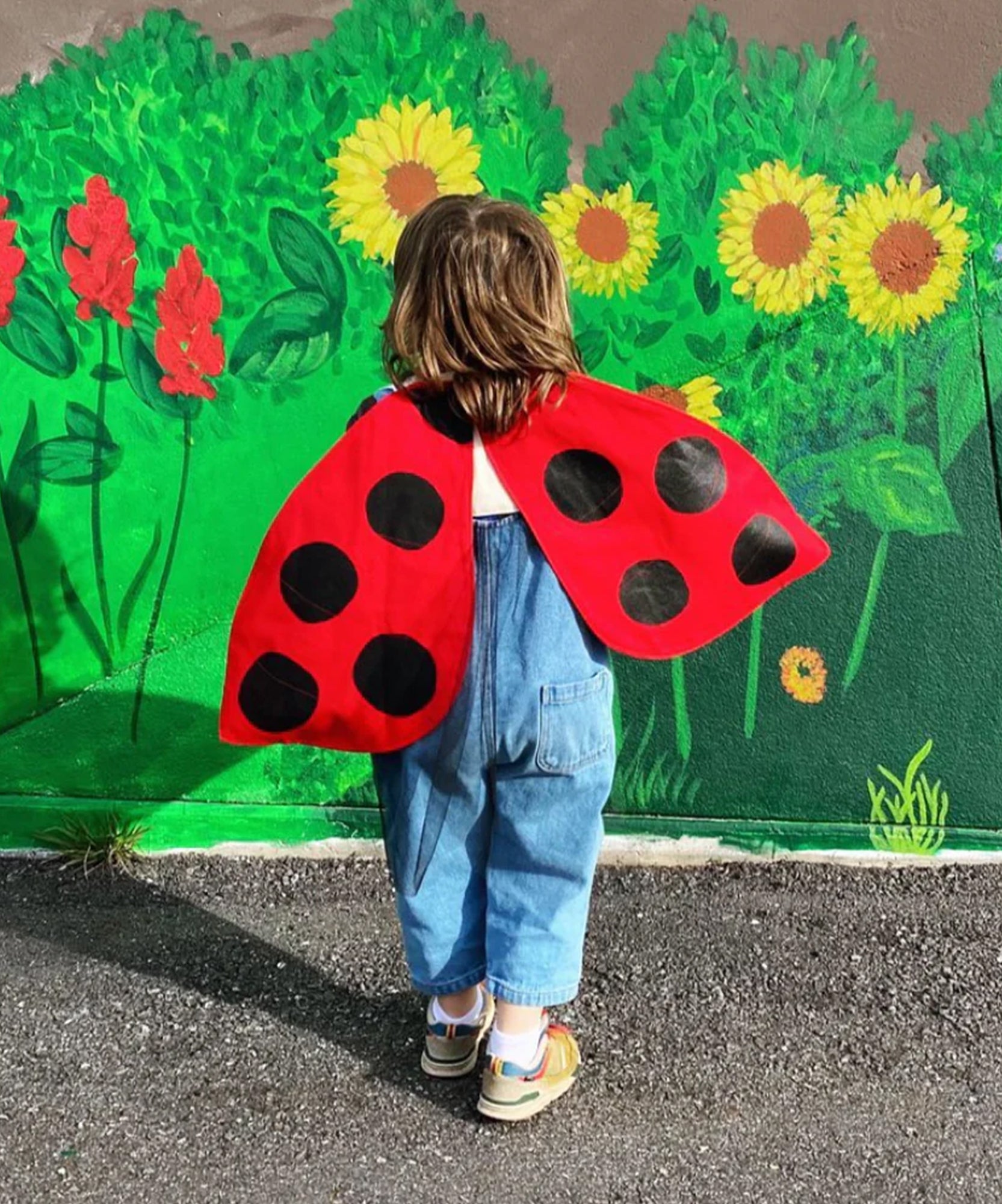 A child wears the Le Ladybug Wings Costume and overalls, standing before a wall painted with green plants and yellow sunflowers.