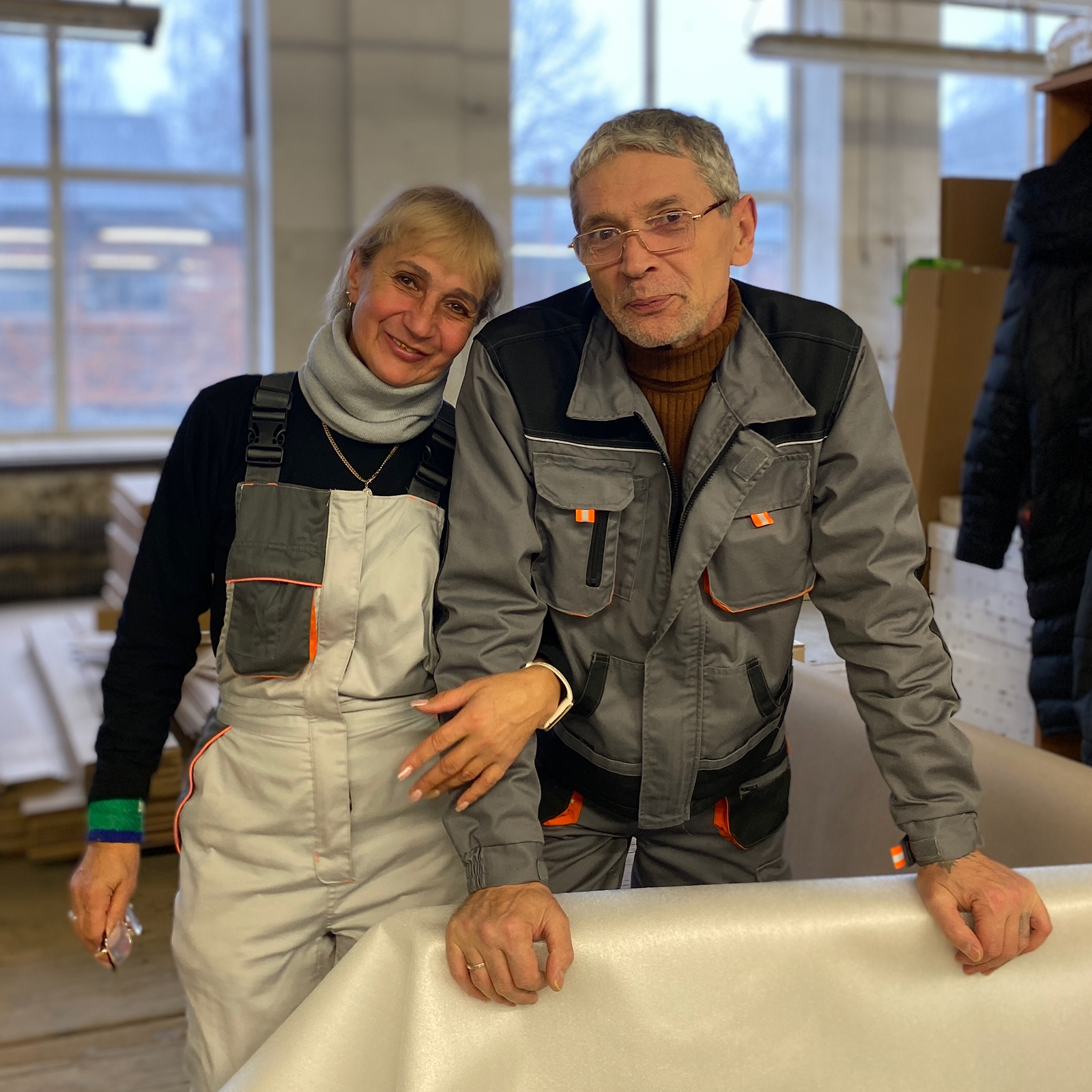 Two people in work uniforms smile in a workshop setting. They stand beside a large piece of white material, with shelves and windows in the background.