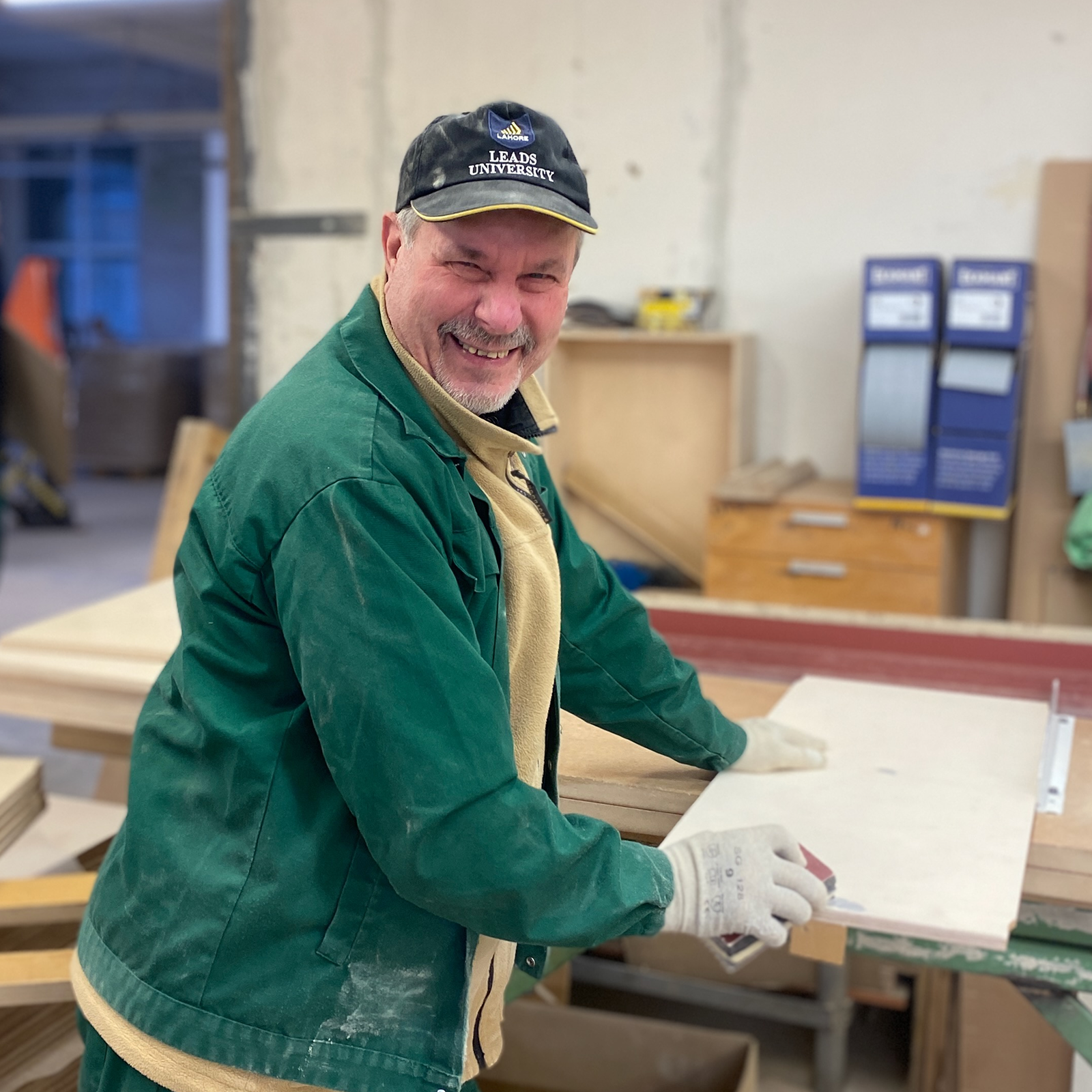 Smiling man in a green jacket and cap works on a wood project in a workshop, using carpentry tools.