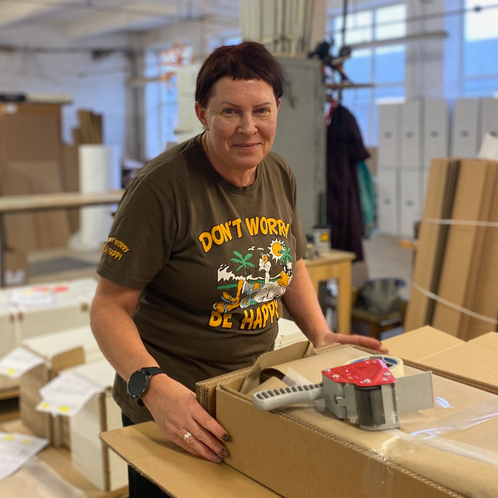 A person in a workshop stands beside an open cardboard box, wearing a Dont Worry Be Happy t-shirt.