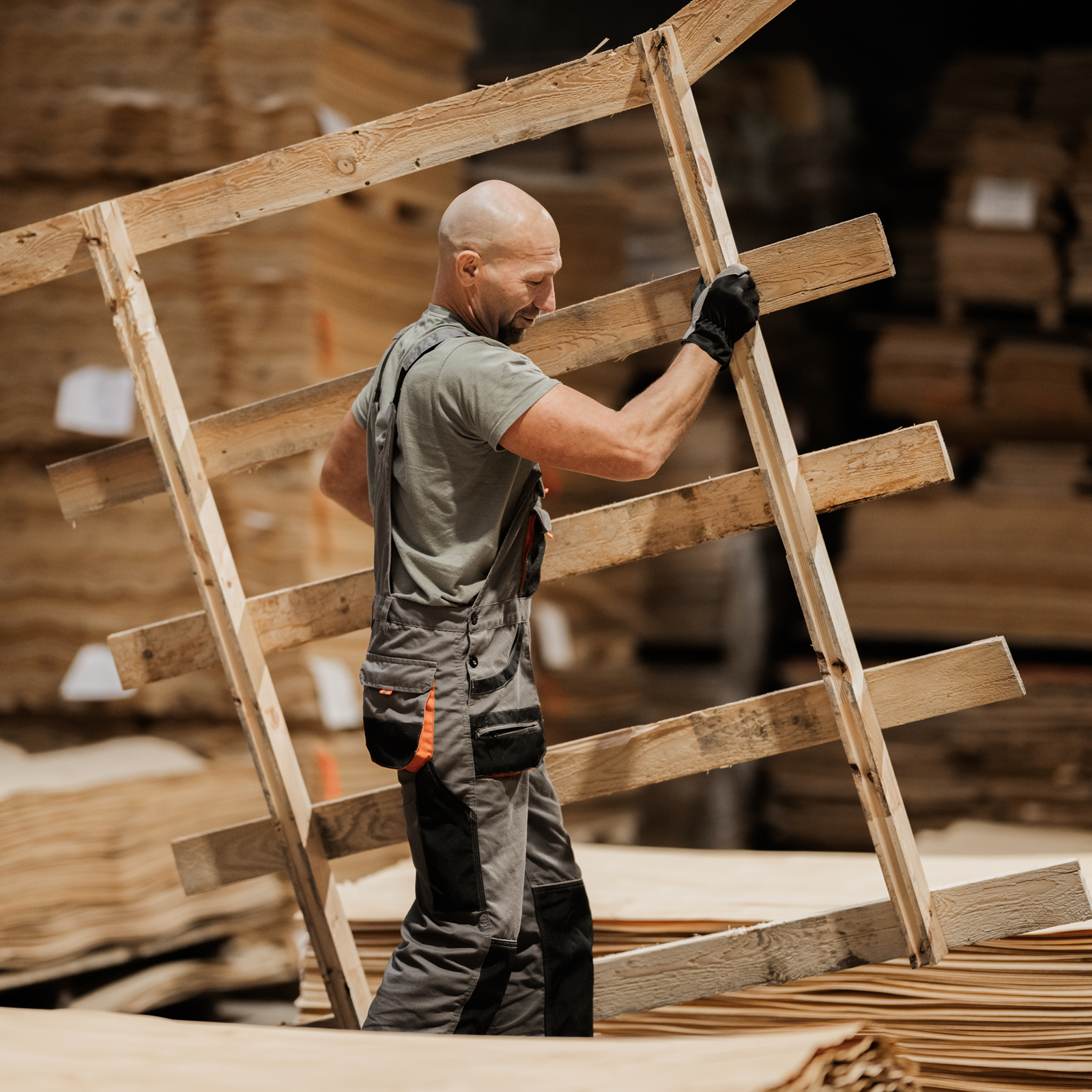 A man in workwear carries a wooden pallet through a lumberyard, with stacks of wood in the background.