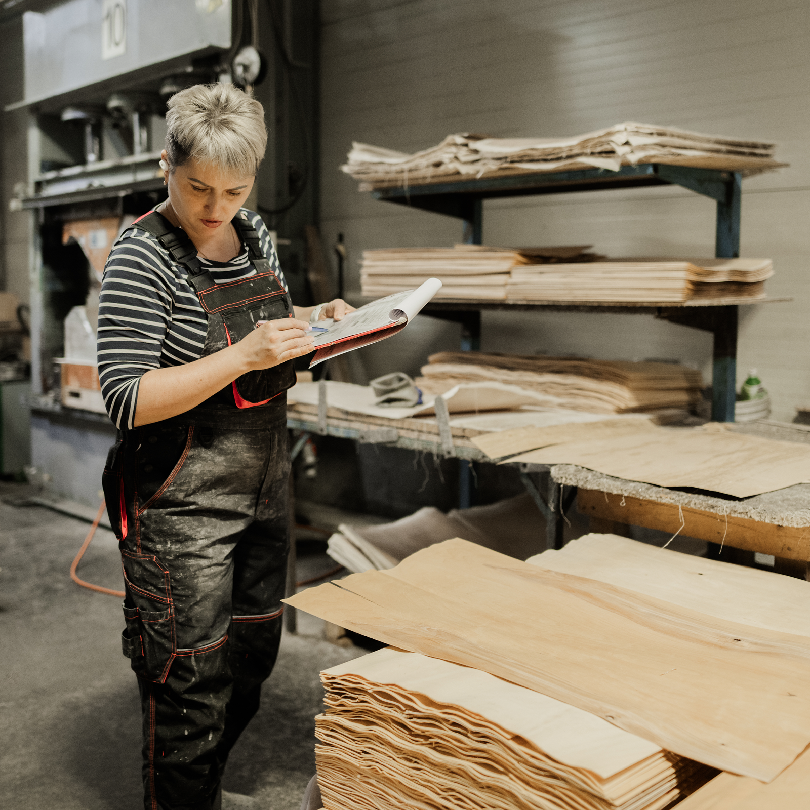 A person in overalls writes on a clipboard while standing next to stacks of wooden sheets in a workshop.