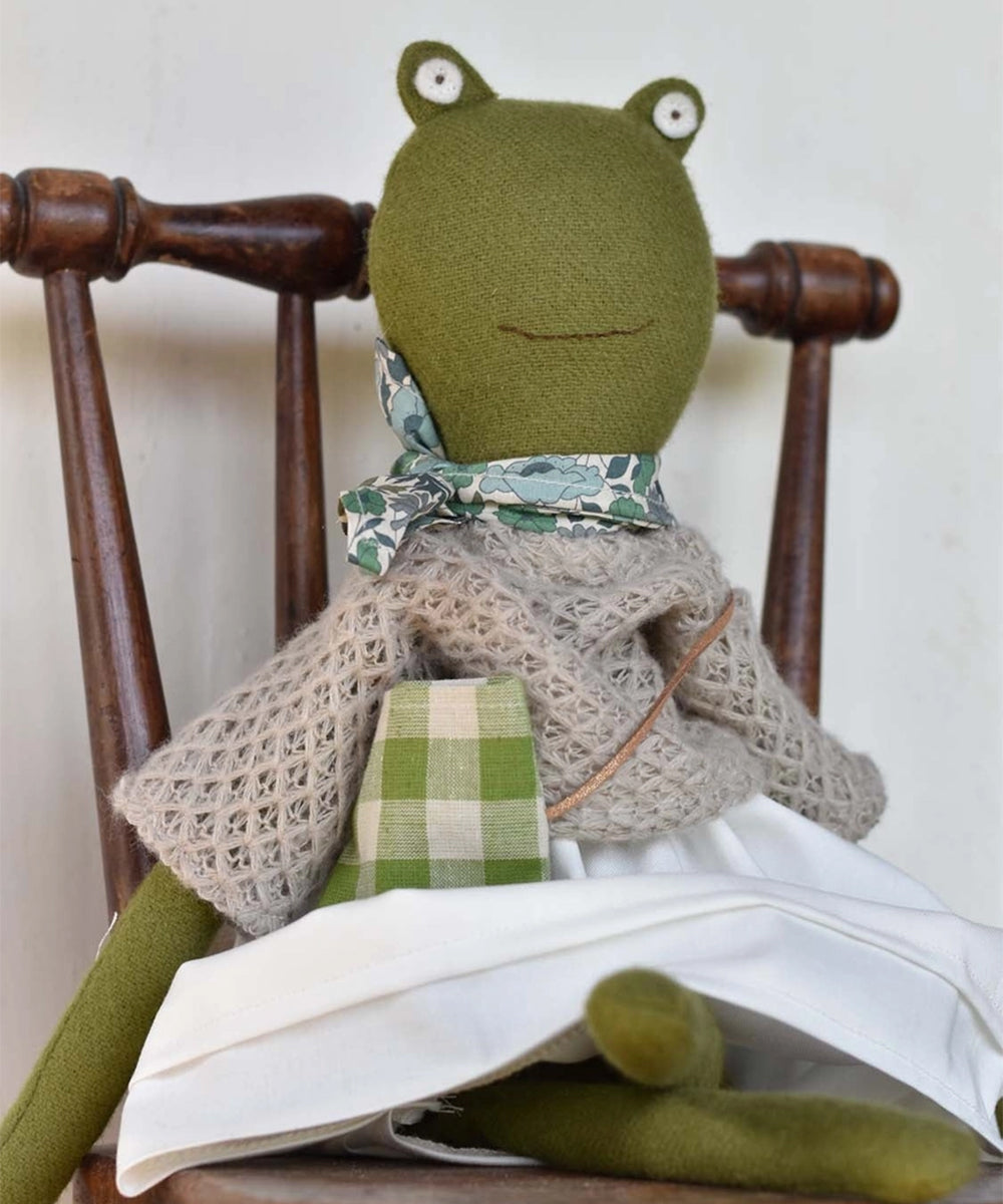 Fern le Frog, wearing a knitted shawl, white skirt, green scarf, and checkered bag, sits on a wooden chair.