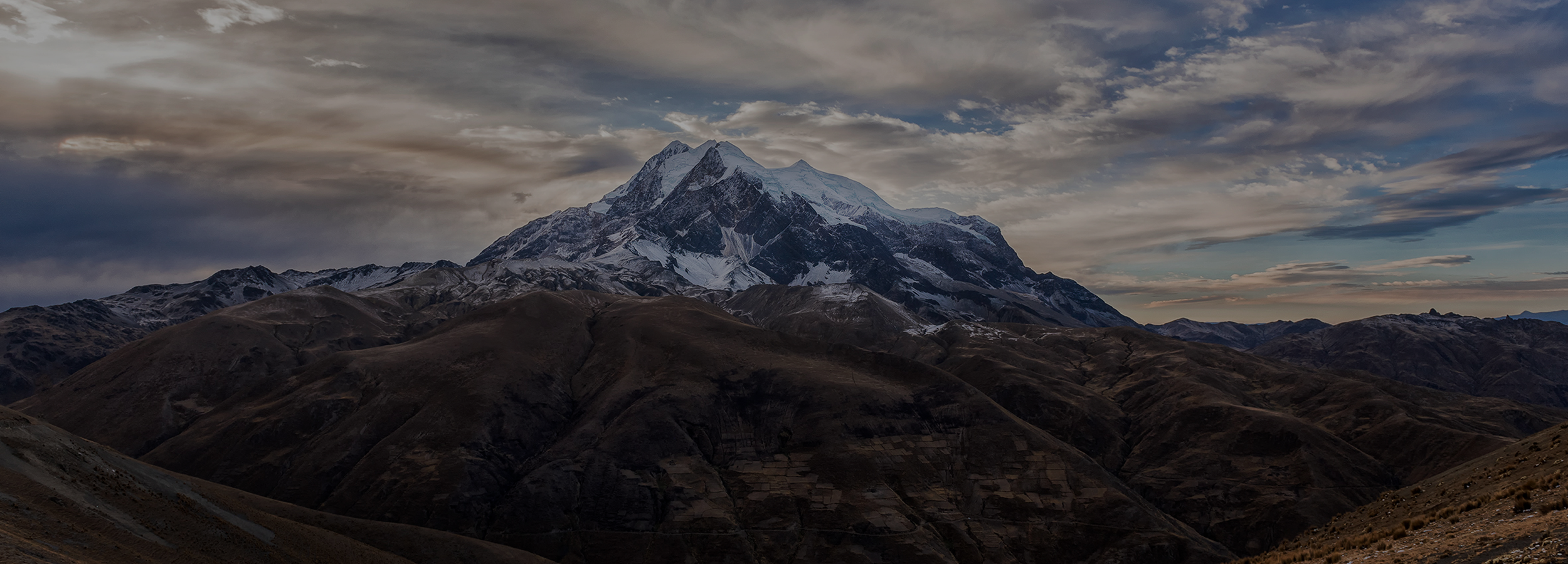 Snow-capped mountain under a dramatic sky with clouds, surrounded by rugged hills and valleys.