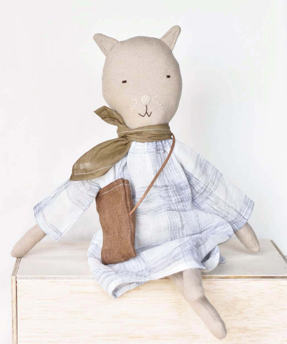 Mae le Cat plush doll wears a plaid dress, green scarf, and brown bag, and sits on a wooden box.