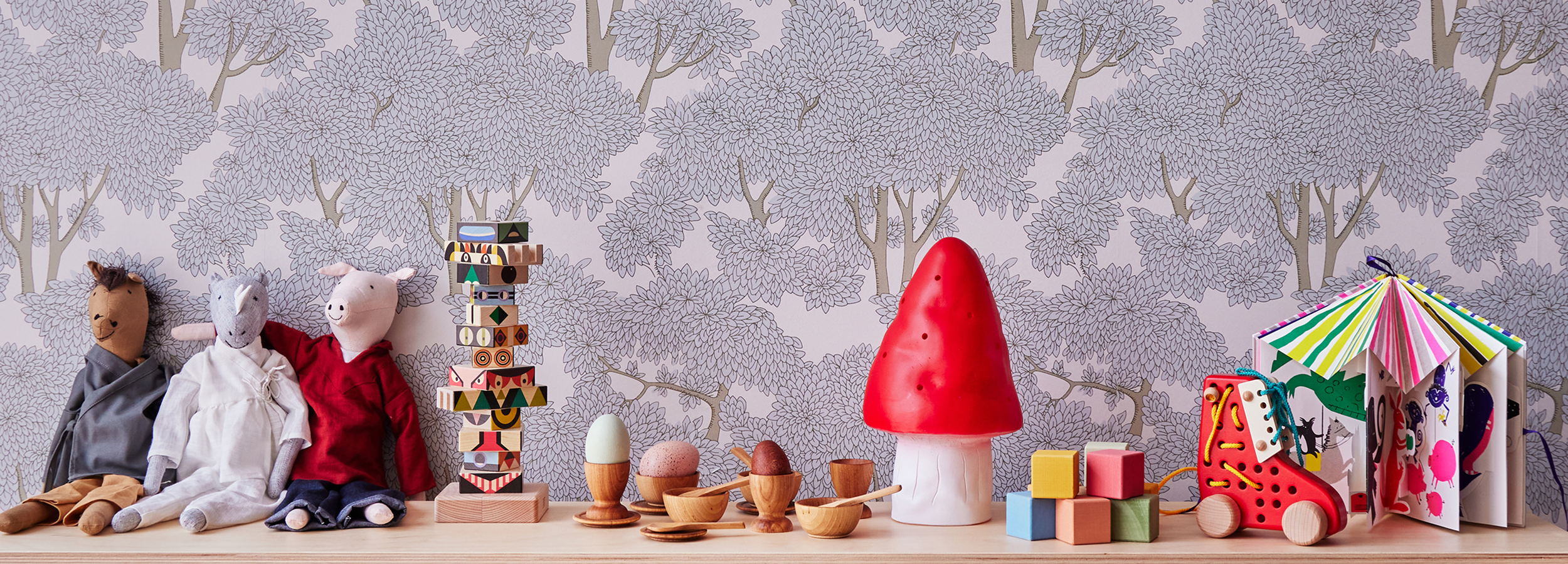 A row of toys on a shelf includes stuffed animals, wooden stacking toys, a red mushroom, wooden bowls, blocks, eggs, and a colorful book in front of patterned tree wallpaper.