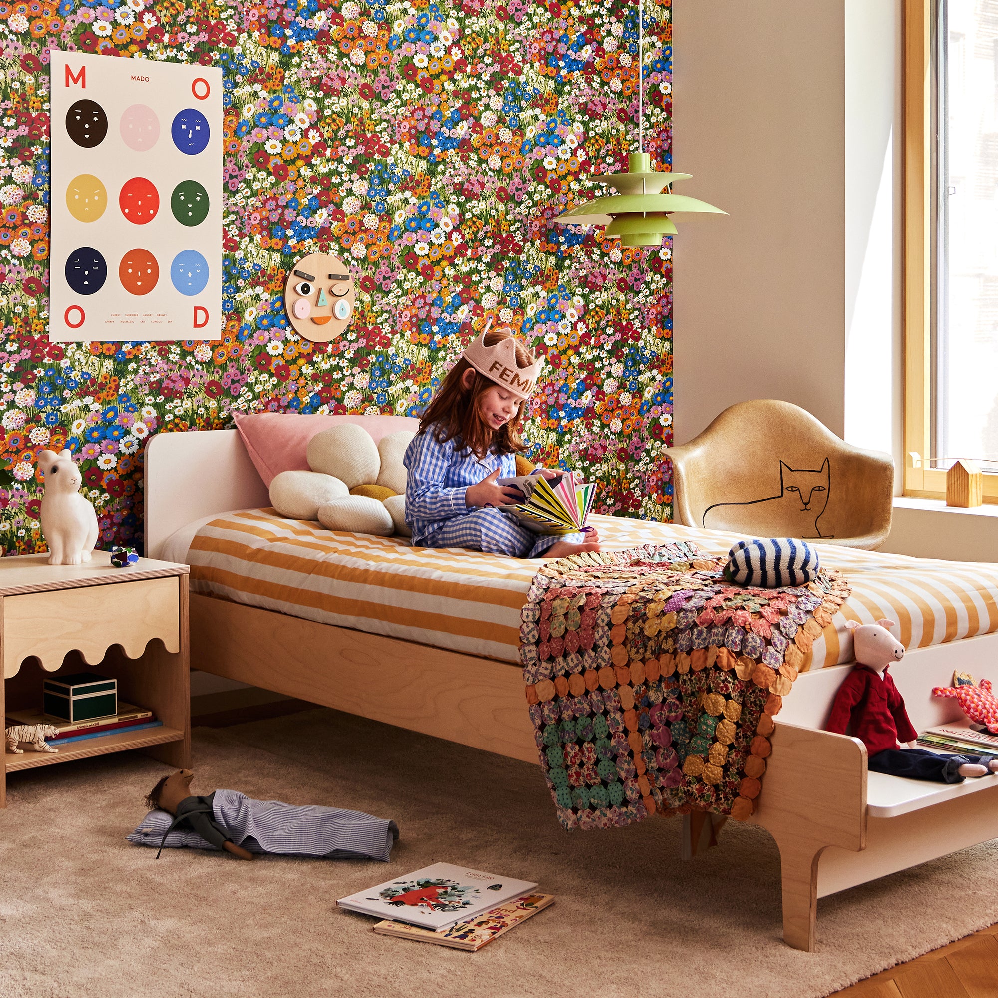 Child sits on a bed in a colorful room with floral wallpaper, reading a book. Toys and a quilt are on the bed. A poster and decorative chair are nearby. Natural light enters through the window.