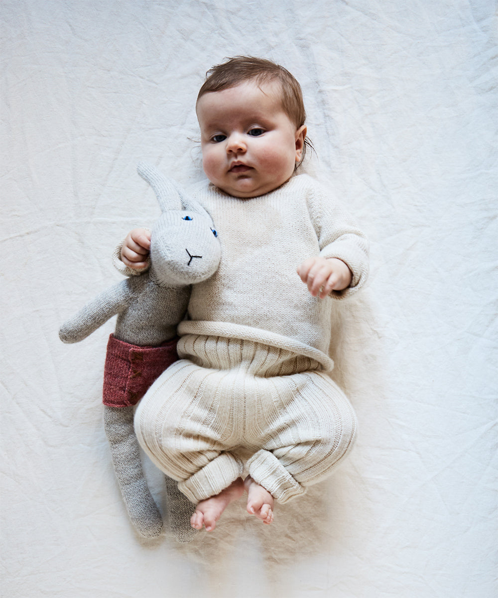 A baby lies on white fabric in a light knit outfit, cozy with a plush gray rabbit featuring a red patch. The serene scene is set atop the Dual Firm Crib Mattress, ensuring comfort and safety with its eco-friendly design.