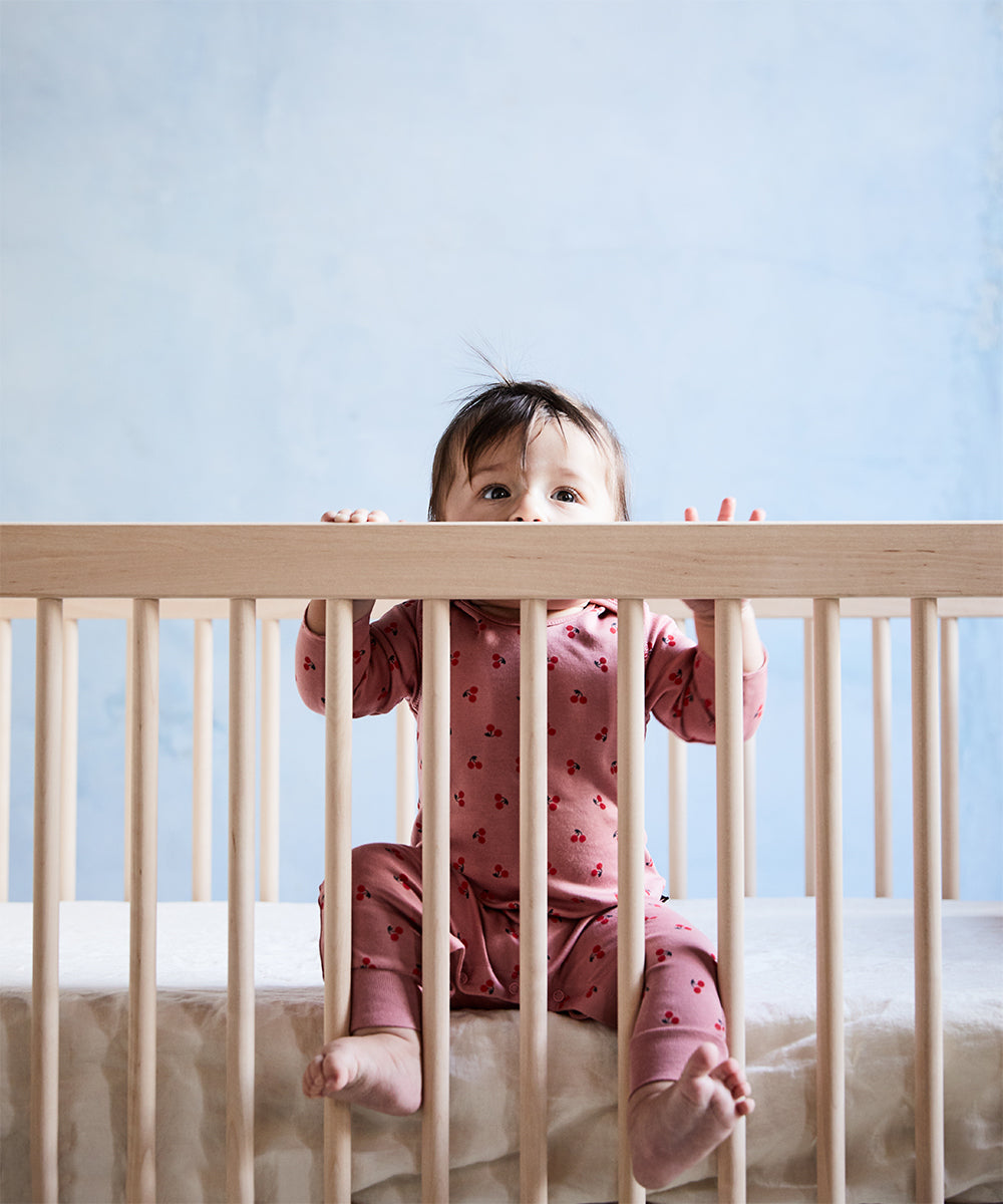 A baby in pink pajamas sits in a wooden crib with a Dual Firm Crib Mattress, curiously peering over the top rail. The background showcases a soft pastel blue wall, creating a serene atmosphere.