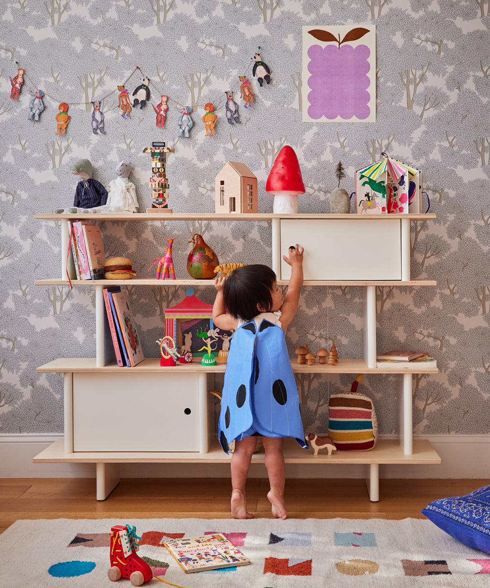 A child in a blue ladybug costume reaches for the Le Wooden Mushroom & Tree Set on a colorful playroom shelf filled with toys and books.