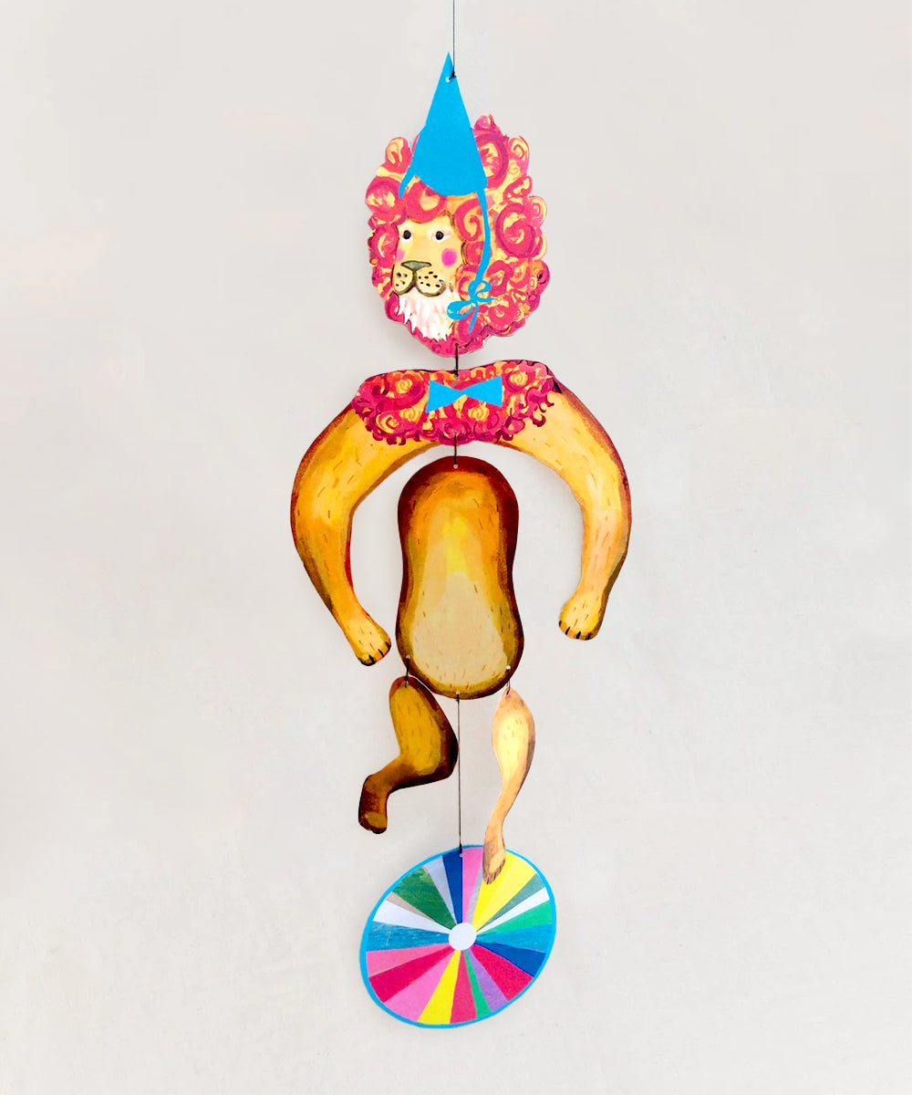 Le Mobile - Circus Lion: A paper lion puppet in a blue hat balances on a colorful wheel against a plain background.