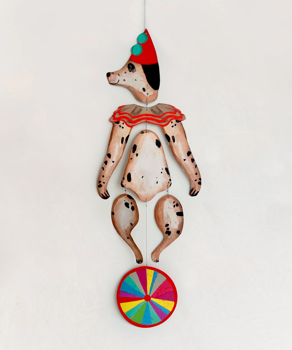 A painted wooden marionette called Le Mobile - Circus Dog, resembling a Dalmatian in a clown outfit with a red hat and multicolored wheel at its feet, hangs against a plain background.