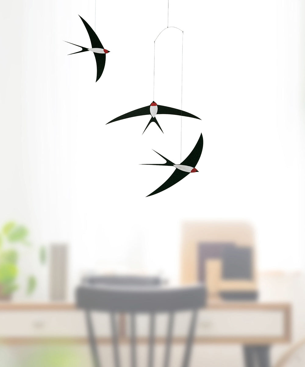 Three Le Mobile - Flying Swallows bird mobiles, in black and white, hang from the ceiling with a blurred chair and desk in the background.