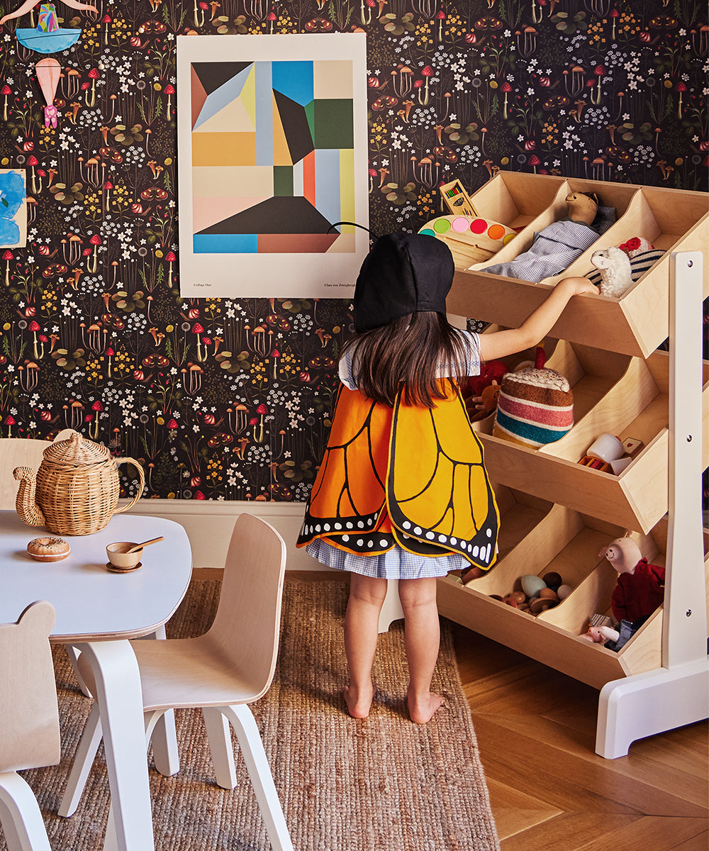 A child wearing Le Monarch Butterfly Wings reaches for toys in a colorful, patterned room with wooden furniture and a wall poster.