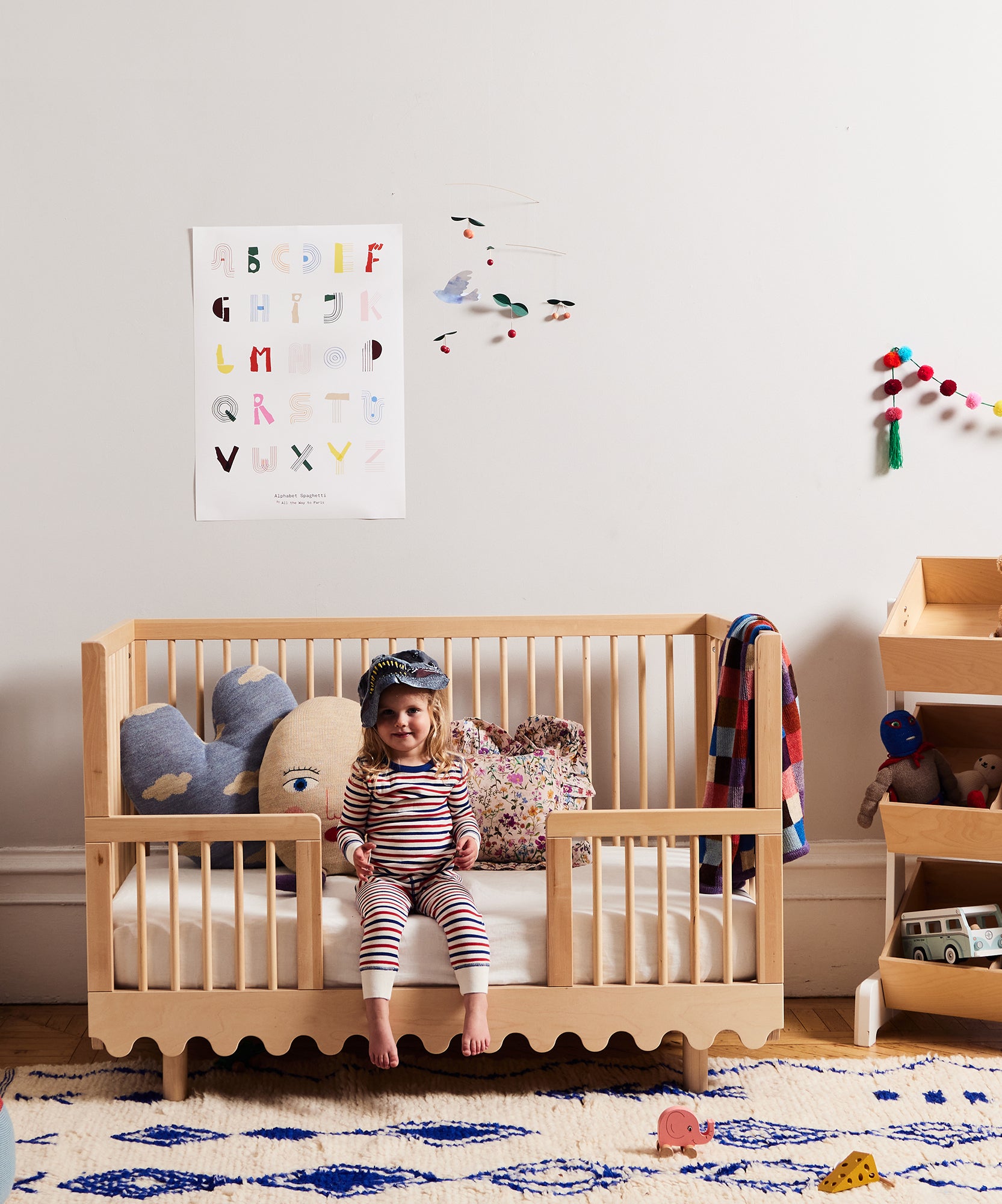 A toddler in striped pajamas sits on a wooden crib converted with the Moss Crib Conversion Kit in a bright, toy-filled nursery.