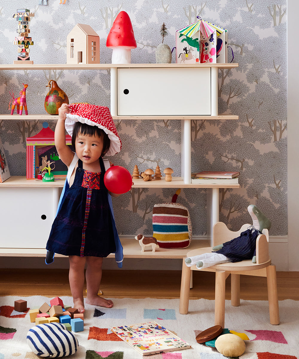 A child in a vibrant room, holding a red ball, wearing a playful hat, surrounded by toys and books, next to the Moss Chair.