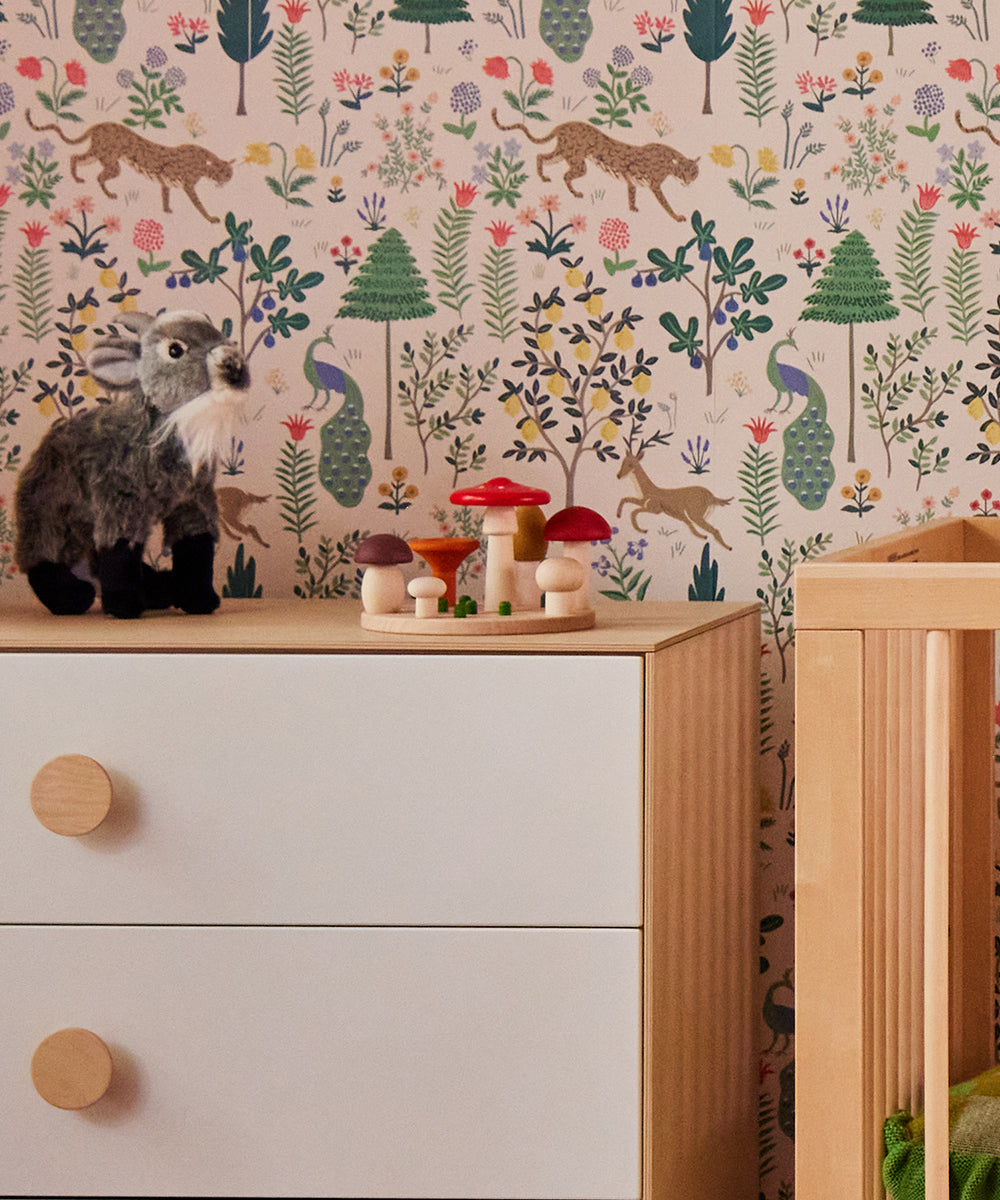 A childrens room with a wooden dresser, Le Mushroom Sorter wooden toys, and a plush toy. The walls are adorned with whimsical forest-themed wallpaper featuring trees, plants, and animals.