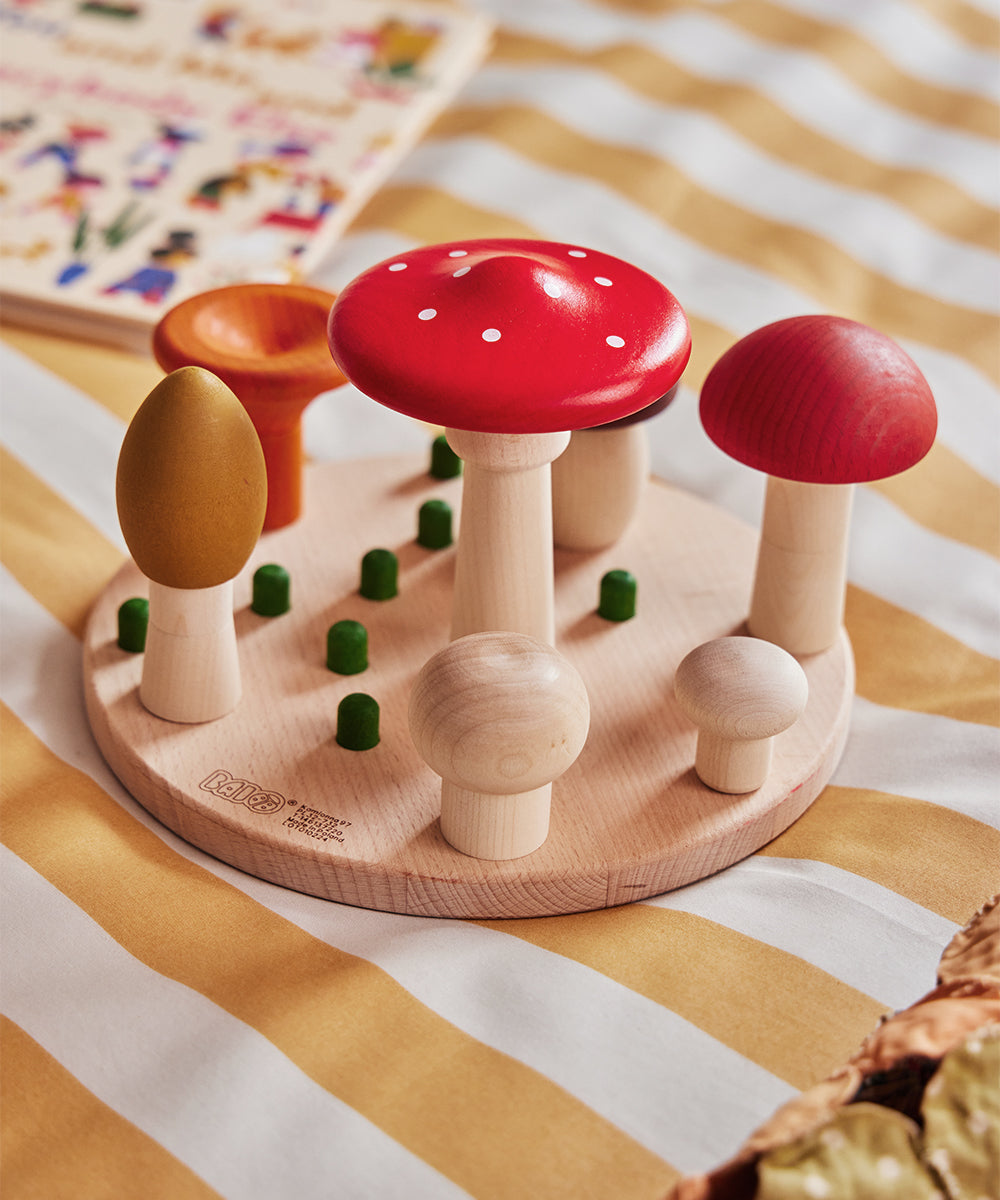 The Le Mushroom Sorter features a variety of wooden toy mushrooms in different sizes and colors, arranged on a round base set against a striped fabric surface.