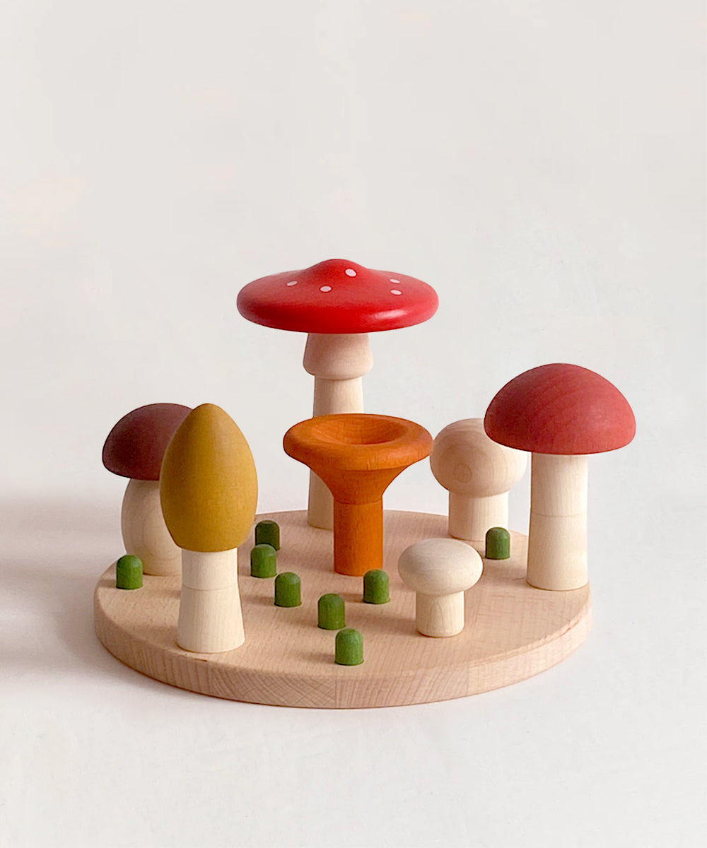 The Le Mushroom Sorter features wooden toy mushrooms in different colors and sizes arranged on a round wooden base, encircled by small green pegs.