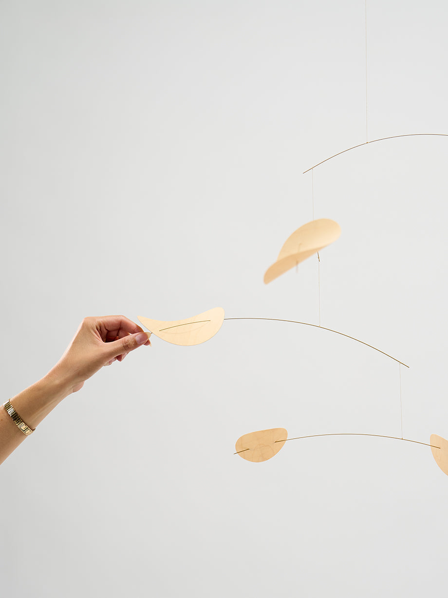 A hand adjusts the Drifting Clouds | Nature | Size M mobile, featuring thin gold rods and simple wooden shapes against a plain background.