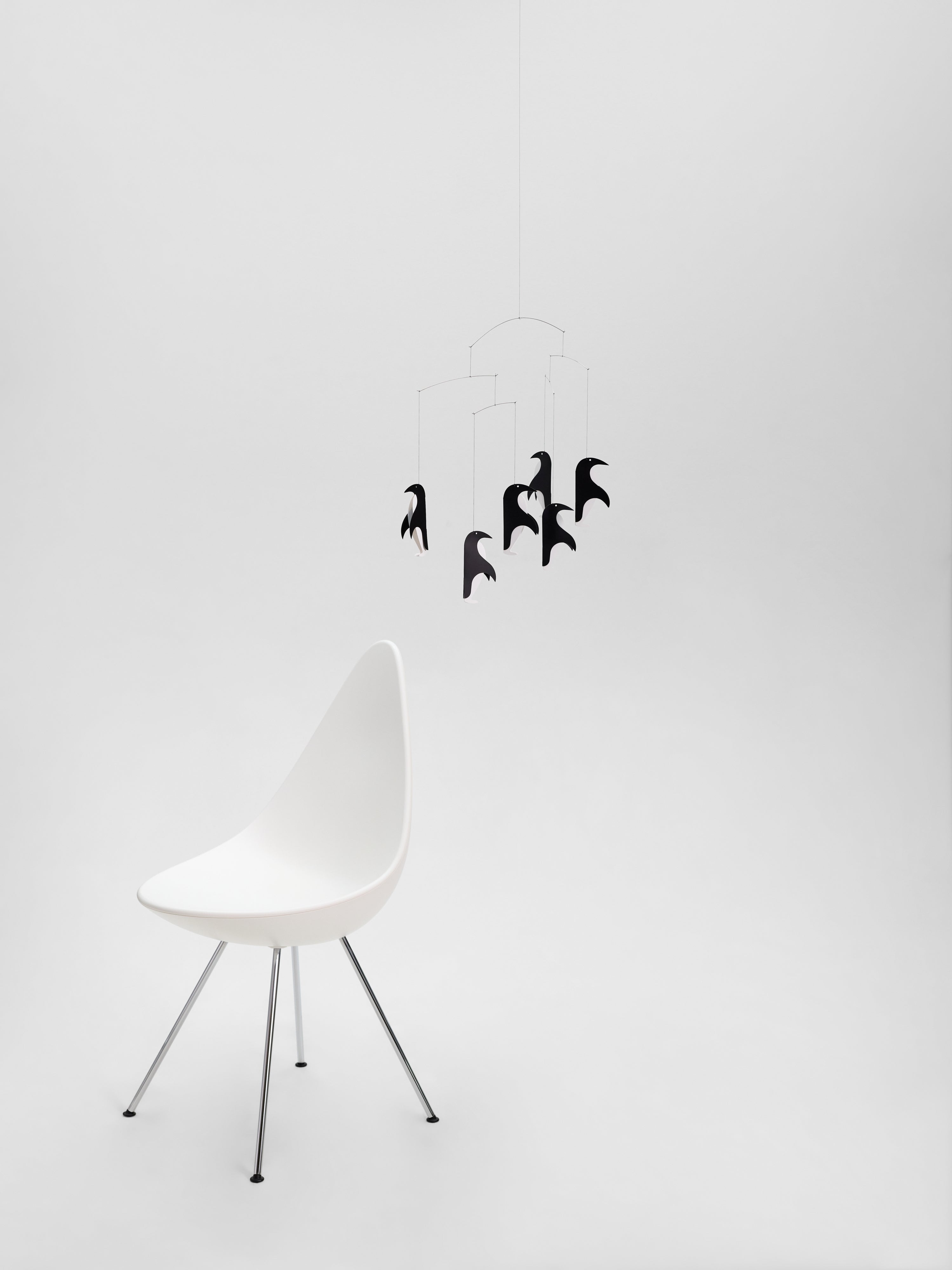 A white modern chair sits next to Penguin Talk, a black bird-shaped hanging mobile, all against a plain white background.