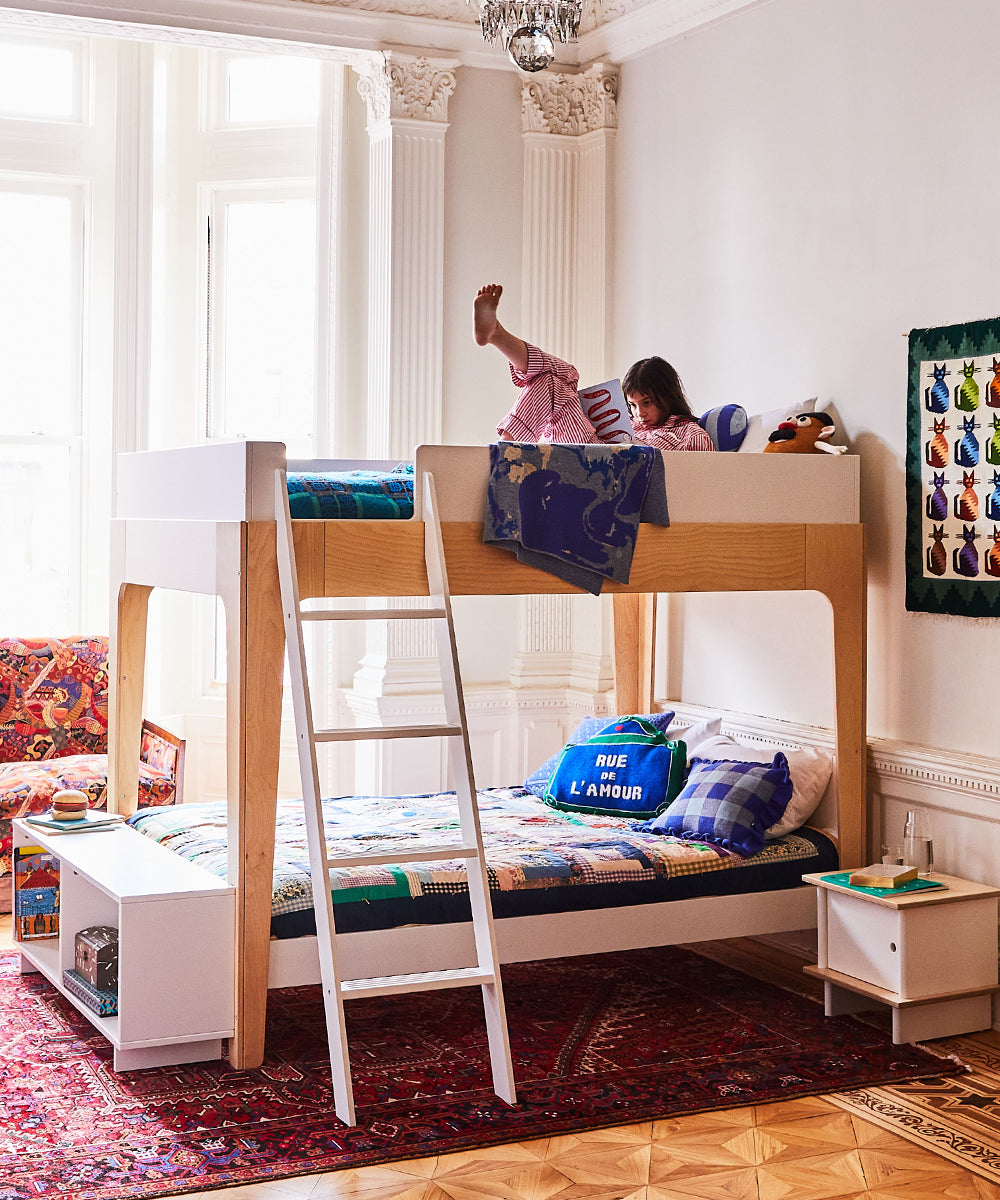 A child in pajamas reads on the top bunk of a Perch Full Bunk Bed, with a neatly made bed below, set in a colorful and cozy room featuring a red rug.