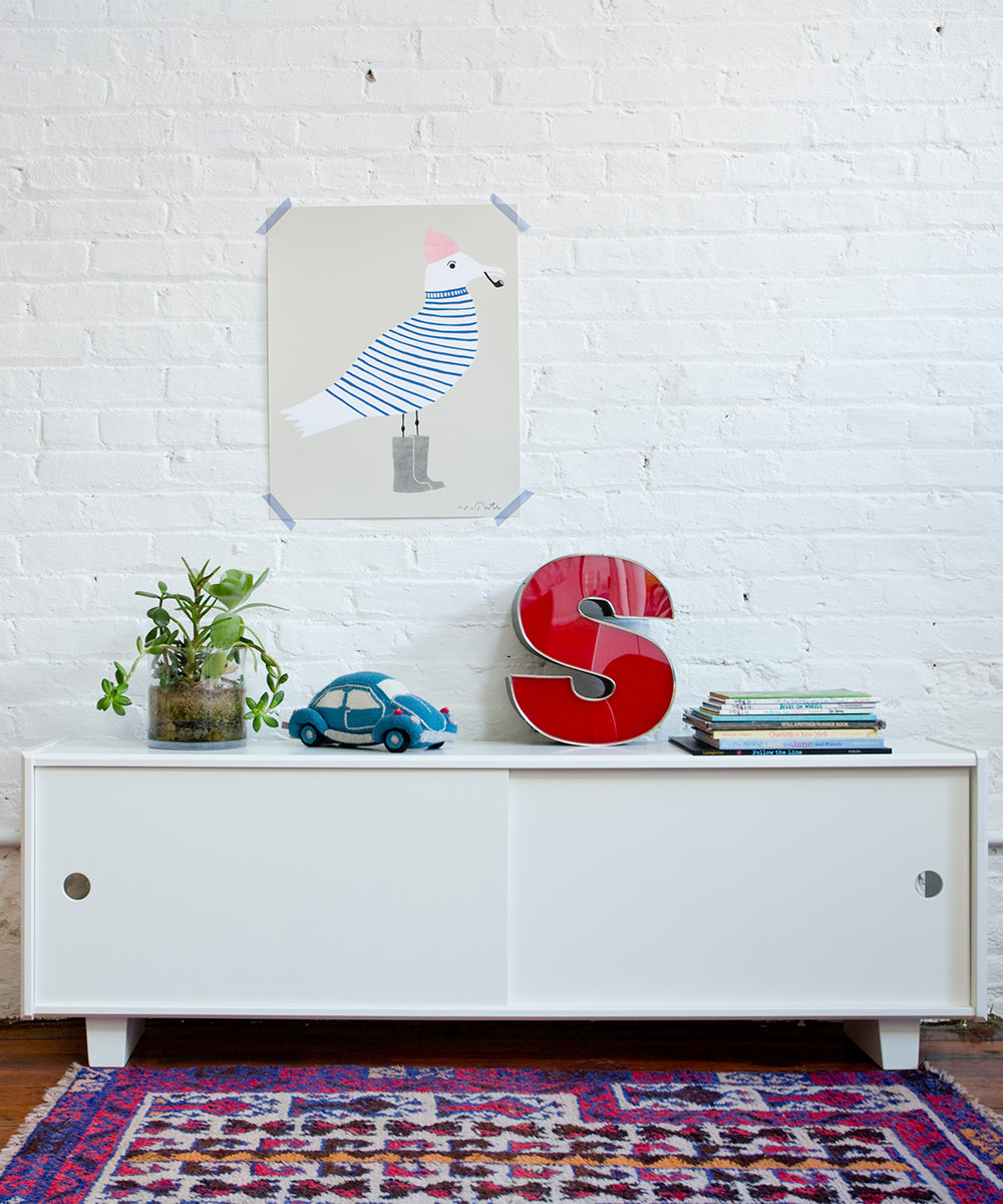 The Perch Console - Full Size in white is styled with plants, a toy car, a red S, and books, set below a bird illustration on a white brick wall.