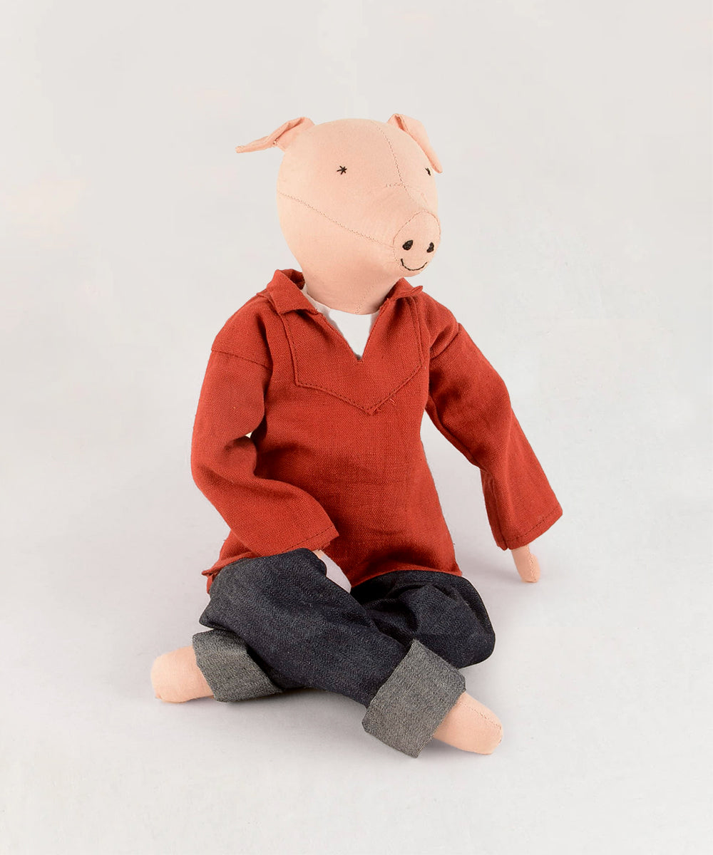 Le Piggy, a stuffed pig toy, wears an orange shirt and blue jeans, sitting on a light grey background.