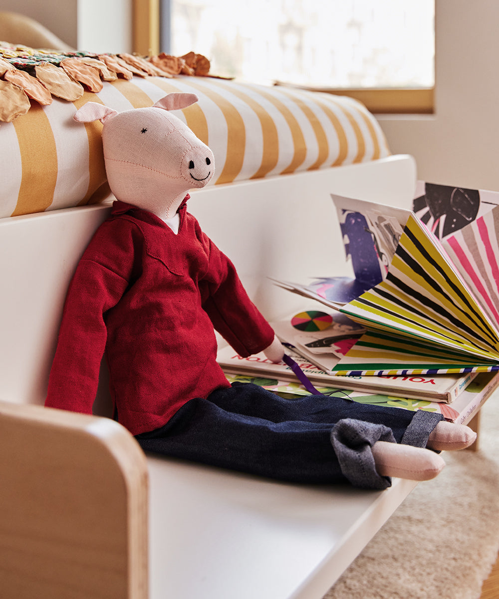 Le Piggy in a red shirt and jeans sits beside open picture books on a child's bed.