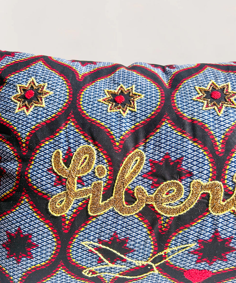 Le Embroidered Pillow - Liberté features gold "Liberté" embroidery with star patterns in red, blue, black, and yellow.