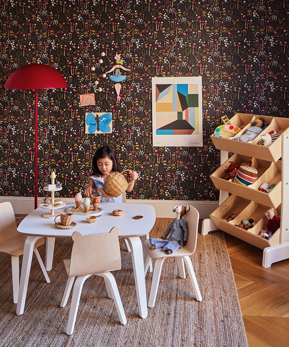 A child plays with Le Doggy toy on a white table in a playroom, featuring patterned wallpaper, a red floor lamp, wall art, and a wooden shelf filled with stuffed animals and toys.